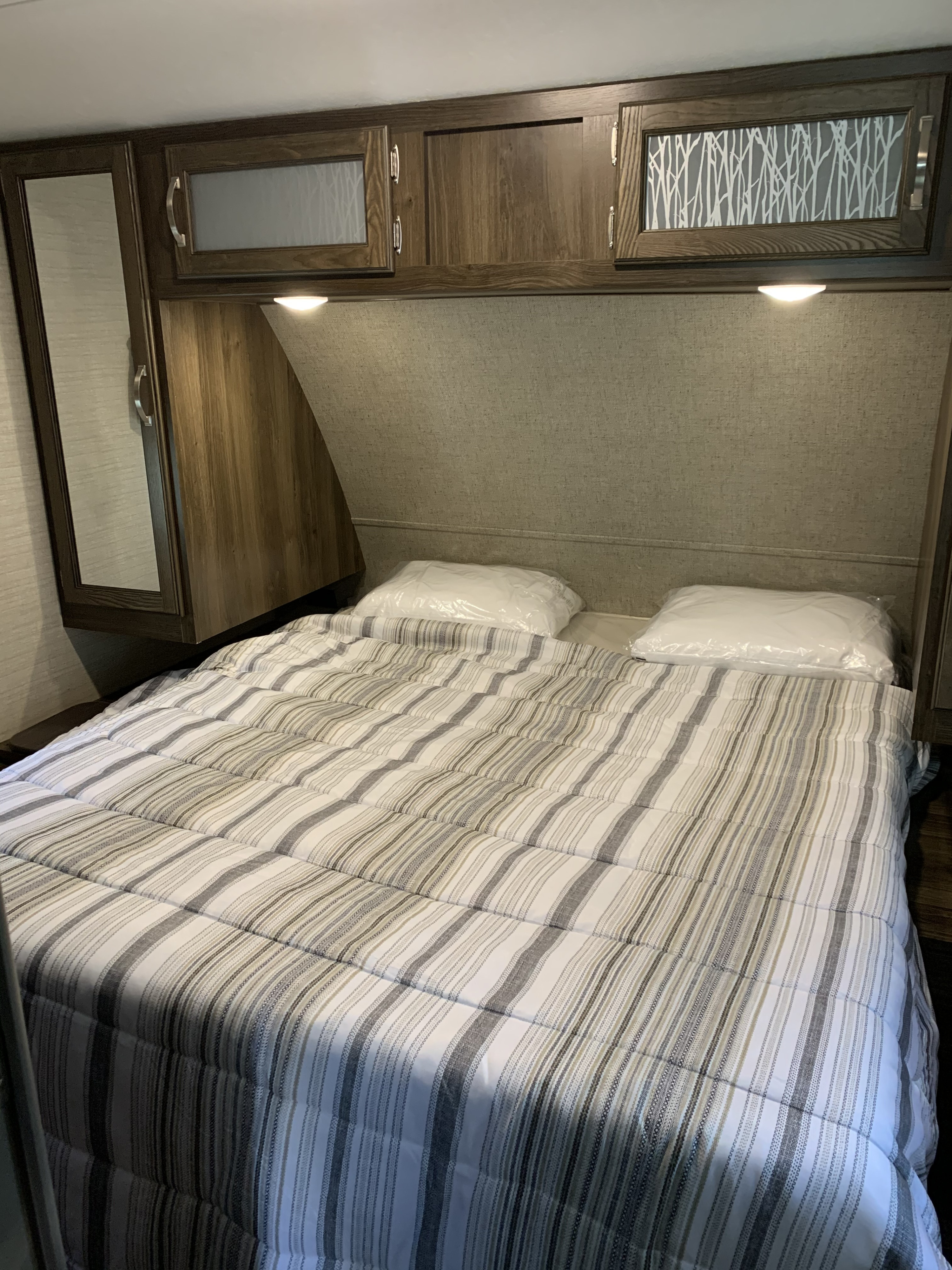 Well-appointed RV rental bedroom features a bed with striped comforter, new pillows, and ample overhead wood cabinets with lighting.. Keystone RV Bullet Ultra Lite 2018