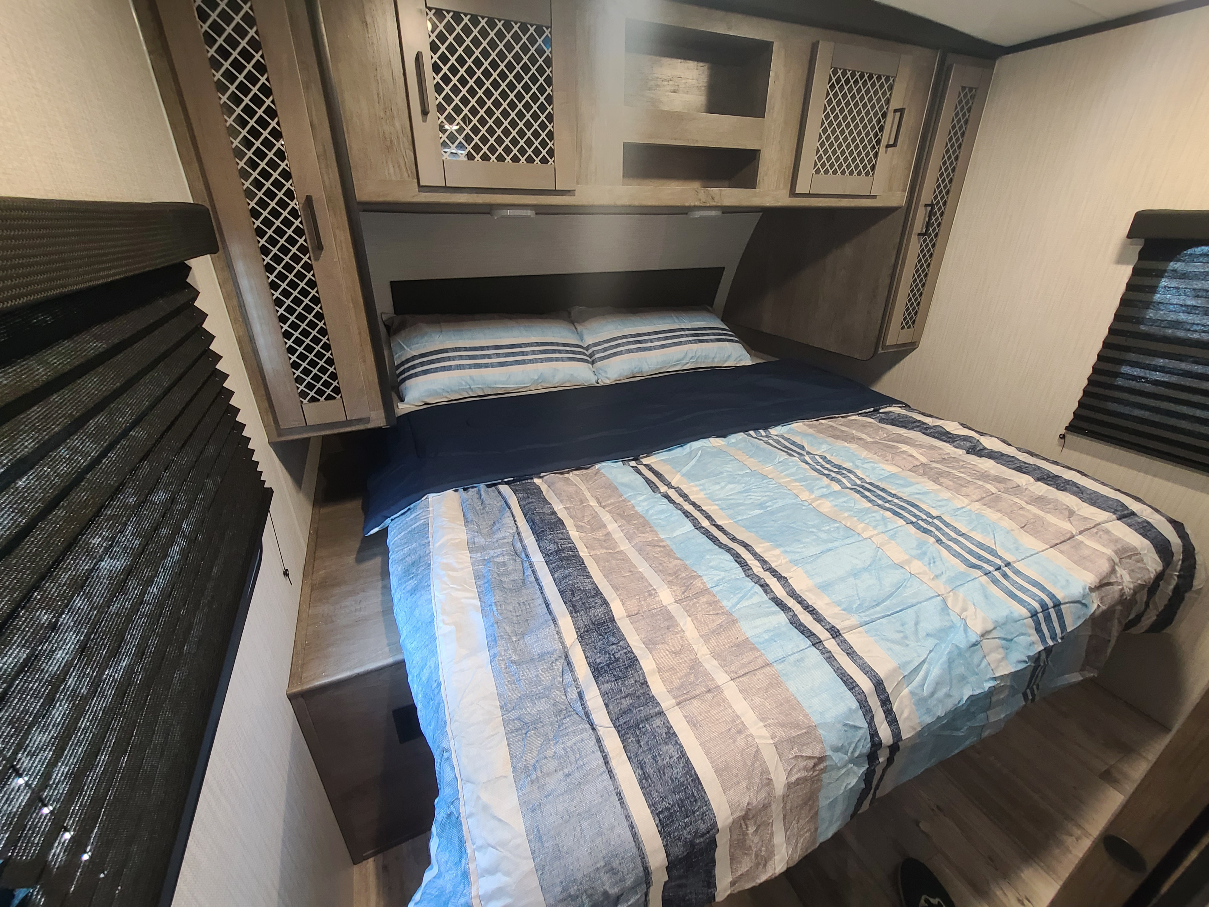 Cozy **RV rental** bedroom with blue striped bedding, overhead cabinets, and dark pleated window blinds for privacy.. Heartland North Trail 24DBS 2021