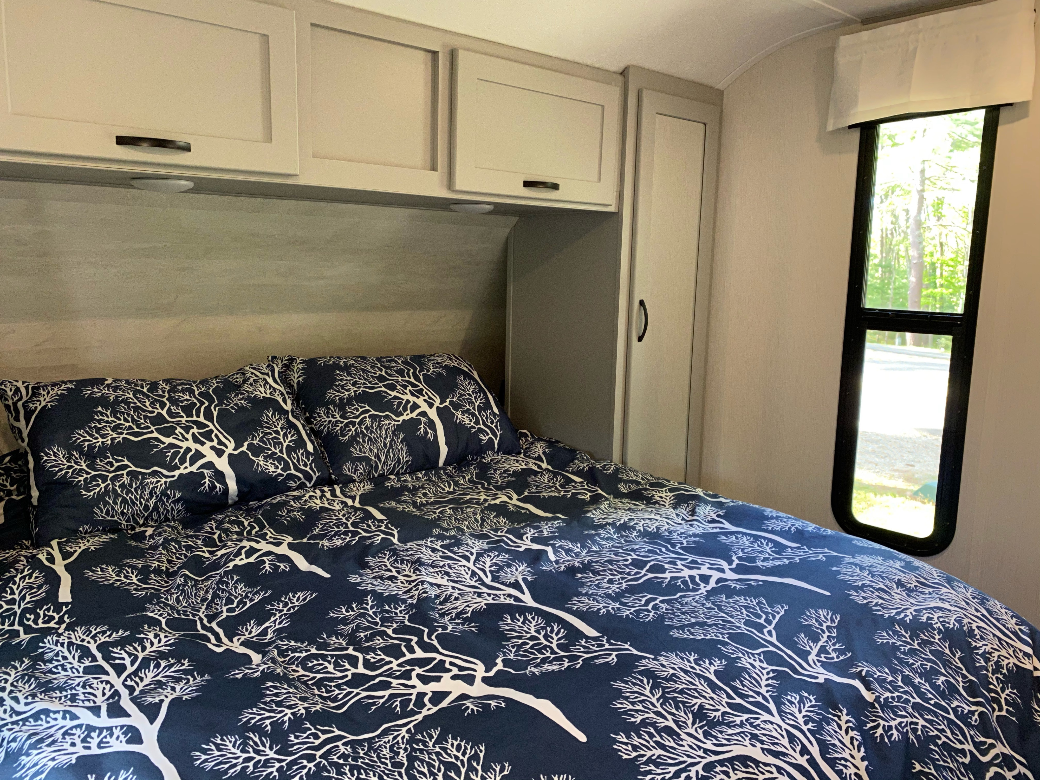 Cozy RV rental bedroom featuring tree-patterned bedding, overhead cabinets, and a window with a nature view.. Forest River Vibe 2021