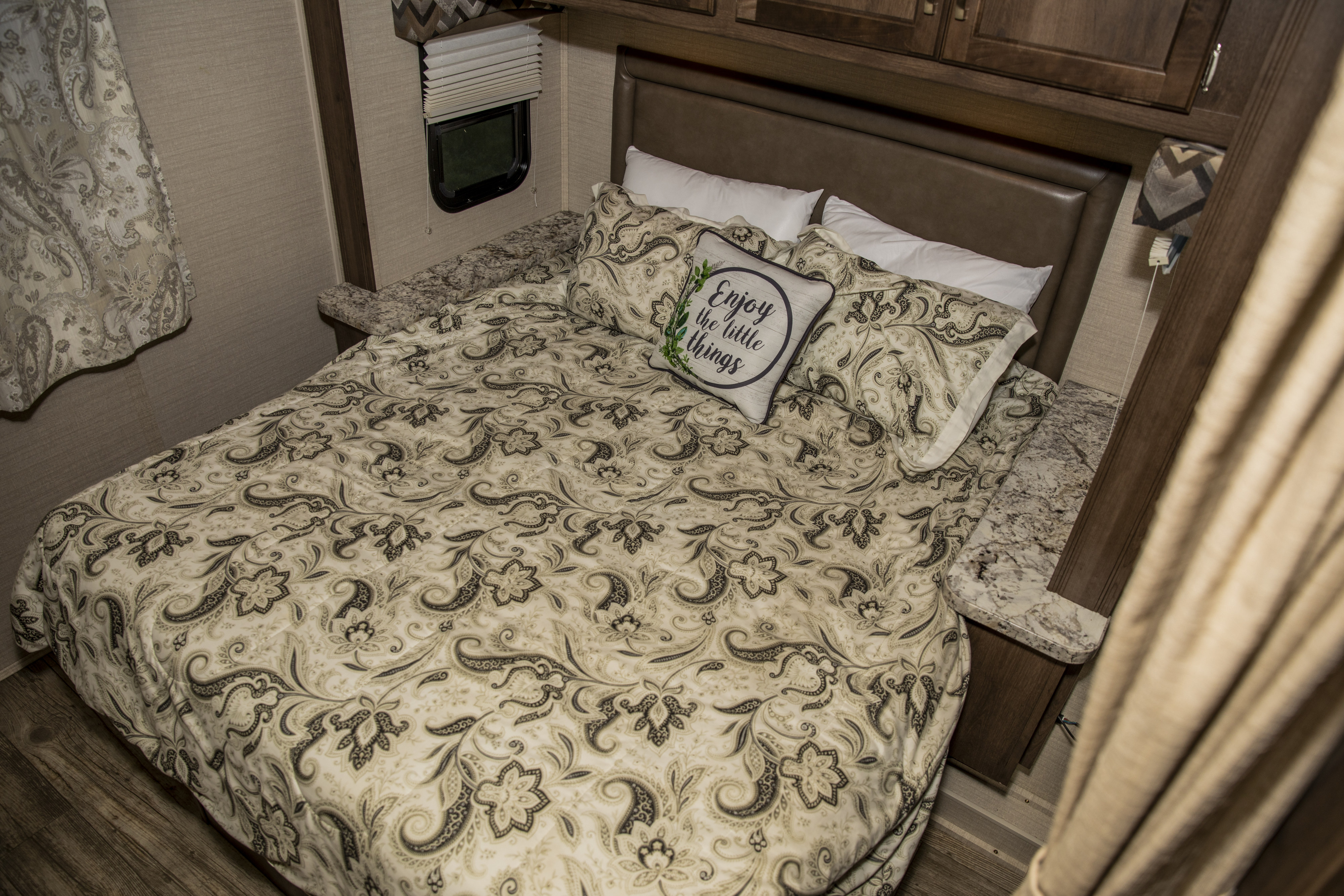 Cozy RV rental bedroom featuring a comfy bed with patterned bedding, a decorative "Enjoy the little things" pillow, and window.. Jayco Redhawk 2018