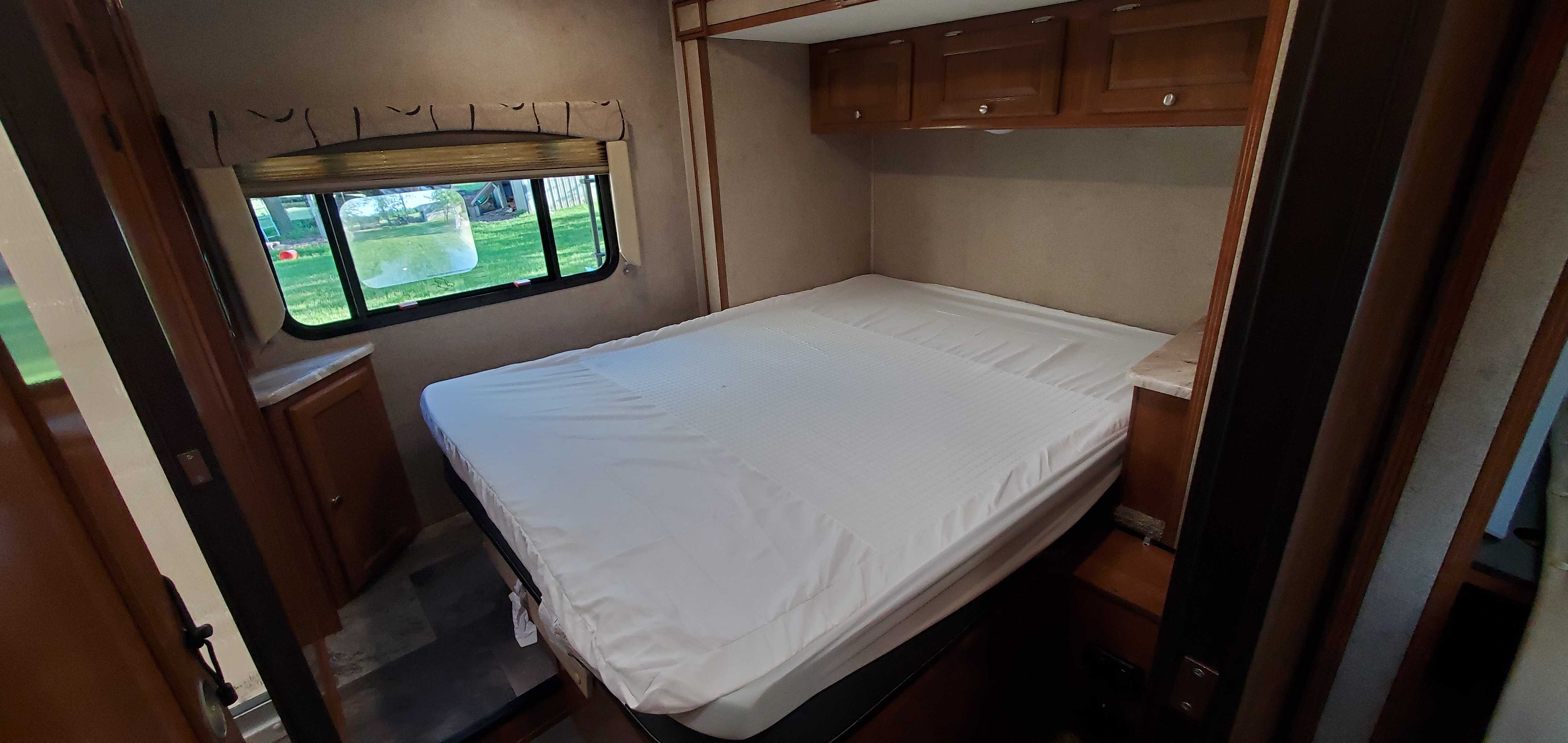 Spacious RV rental bedroom featuring a queen bed with white protector, overhead cabinets, and large window.. Coachmen Leprechaun 2019