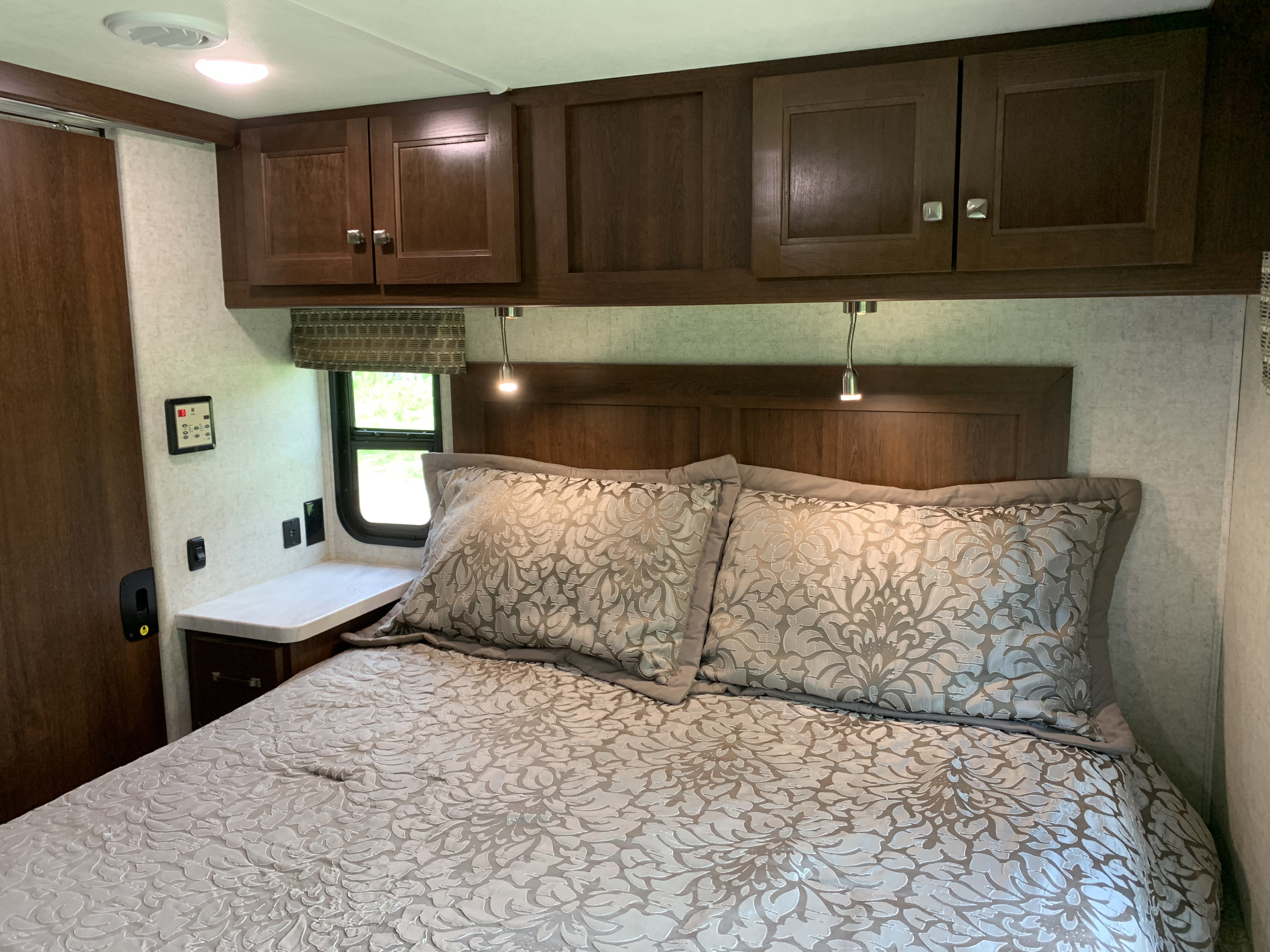 Interior of an RV rental bedroom showcasing a large bed with patterned bedding, dark wood cabinets, and functional reading lights.. Forest River Sunseeker 2019