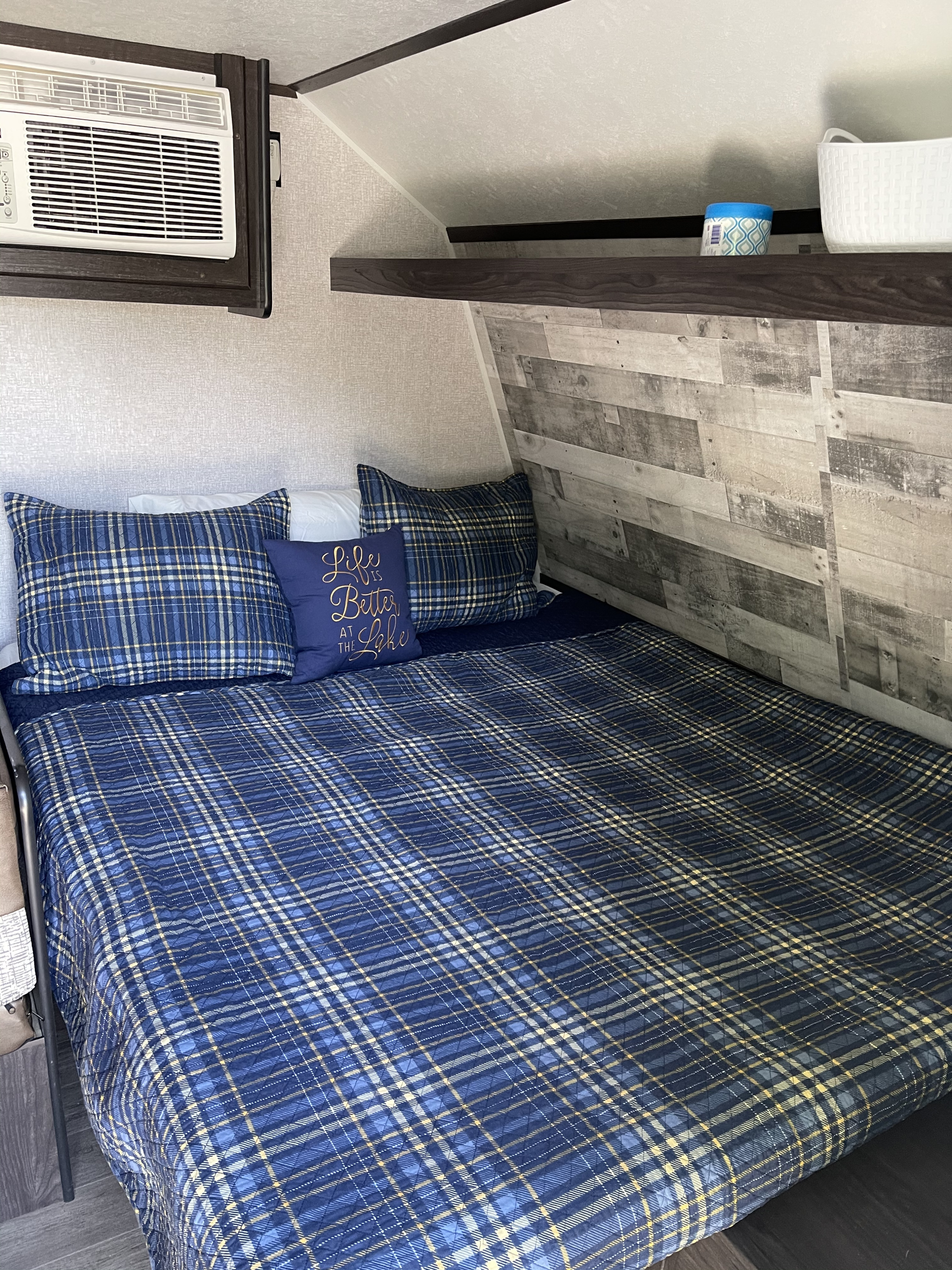 Cozy RV rental bedroom features a blue plaid bed with 'lake' pillow, an AC unit, and wood accents.. Dutchmen Coleman 2021