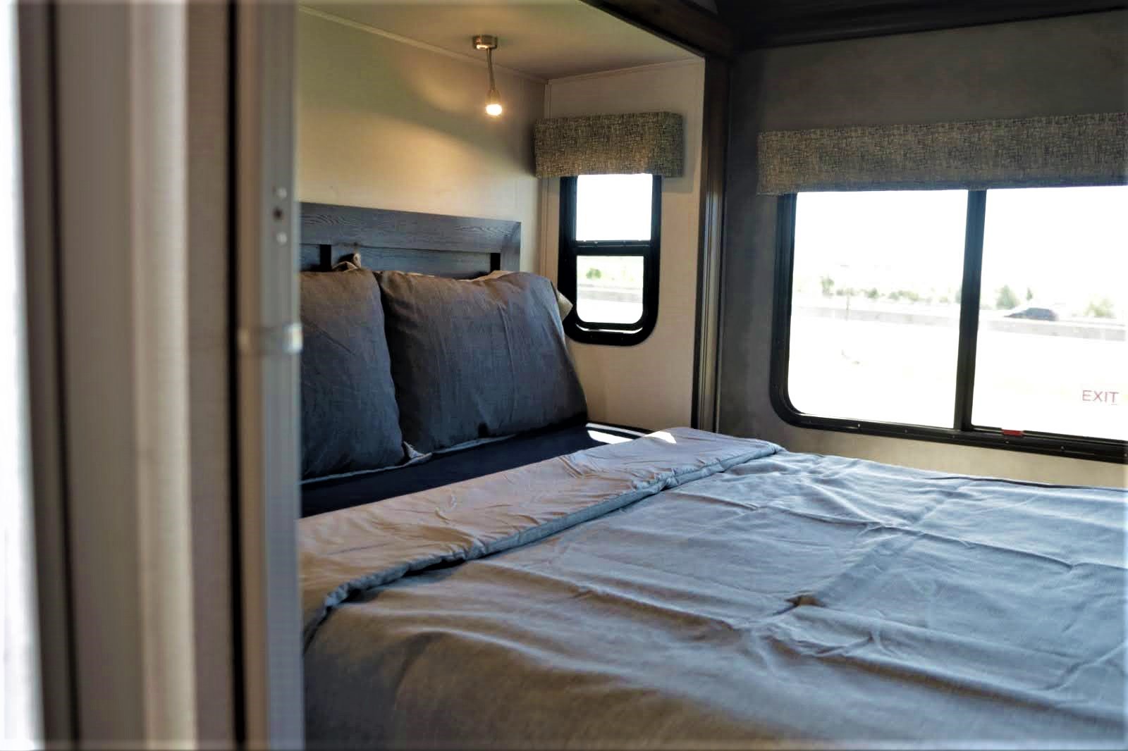 Cozy RV rental bedroom with a dark wood headboard, grey bed, and large windows offering bright outdoor views.. Forest River Other 2021