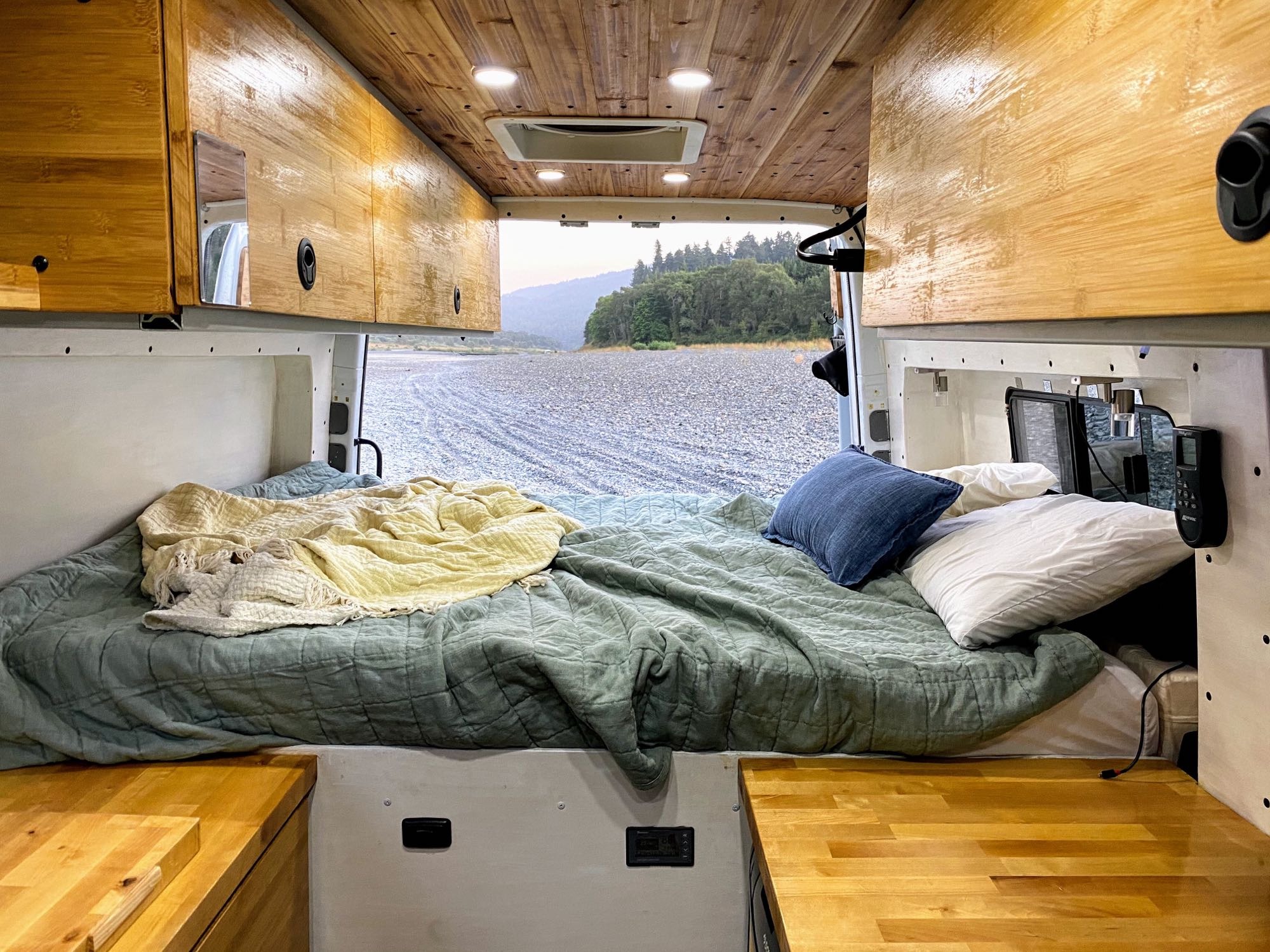 Inside an rv rental camper van: a cozy bed with green blankets, wooden interiors, and a view of a rocky riverbed and mountains.. Mercedes-Benz Sprinter 2018