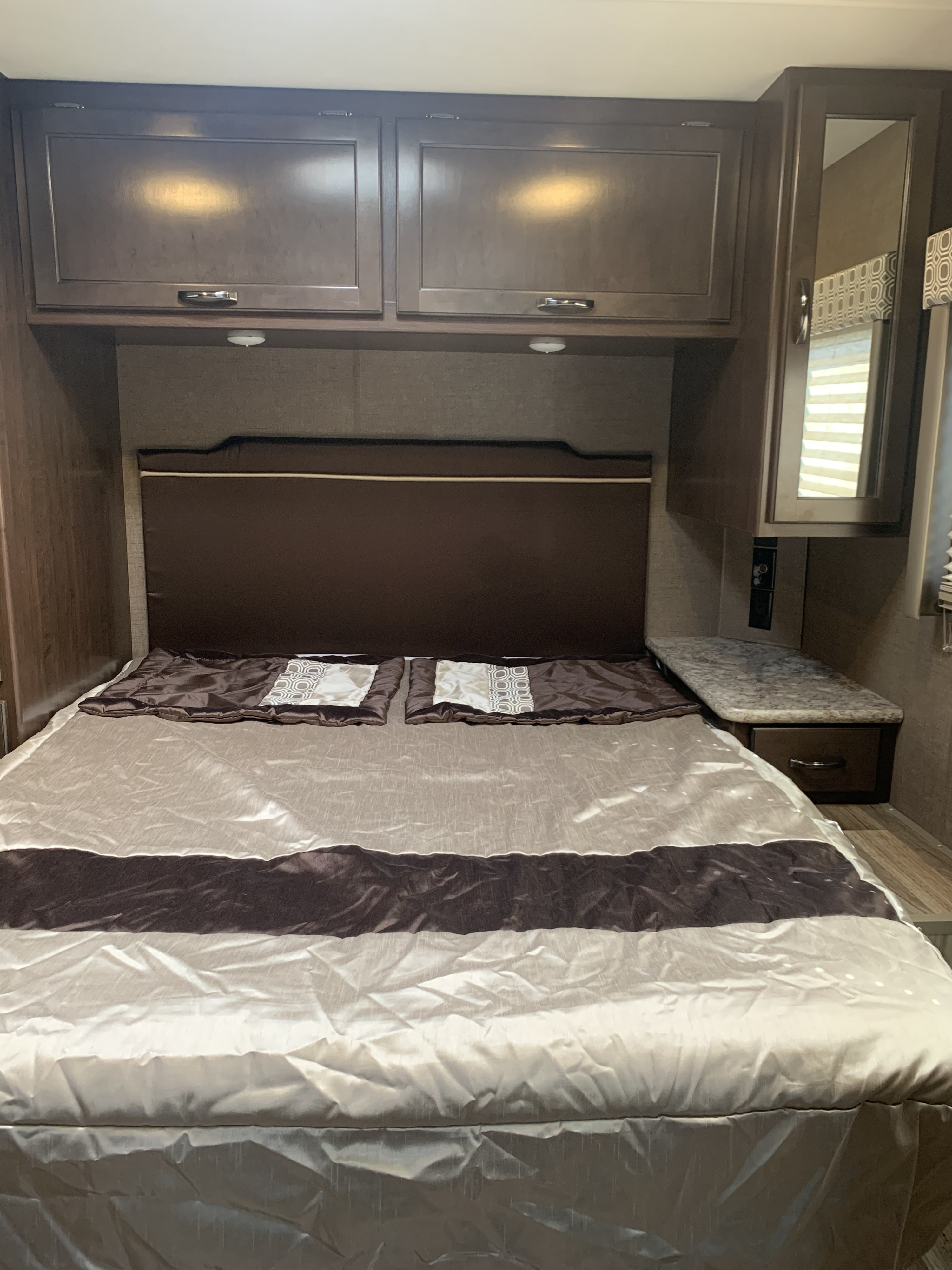 Cozy RV rental bedroom features a comfortable bed with brown and beige bedding, overhead cabinets, and a nightstand.. Thor Motor Coach Four Winds 2019