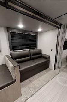 This RV rental interior shows a dark sofa, dinette, and window in a modern living space with light walls and flooring.. Keystone Hideout 2021