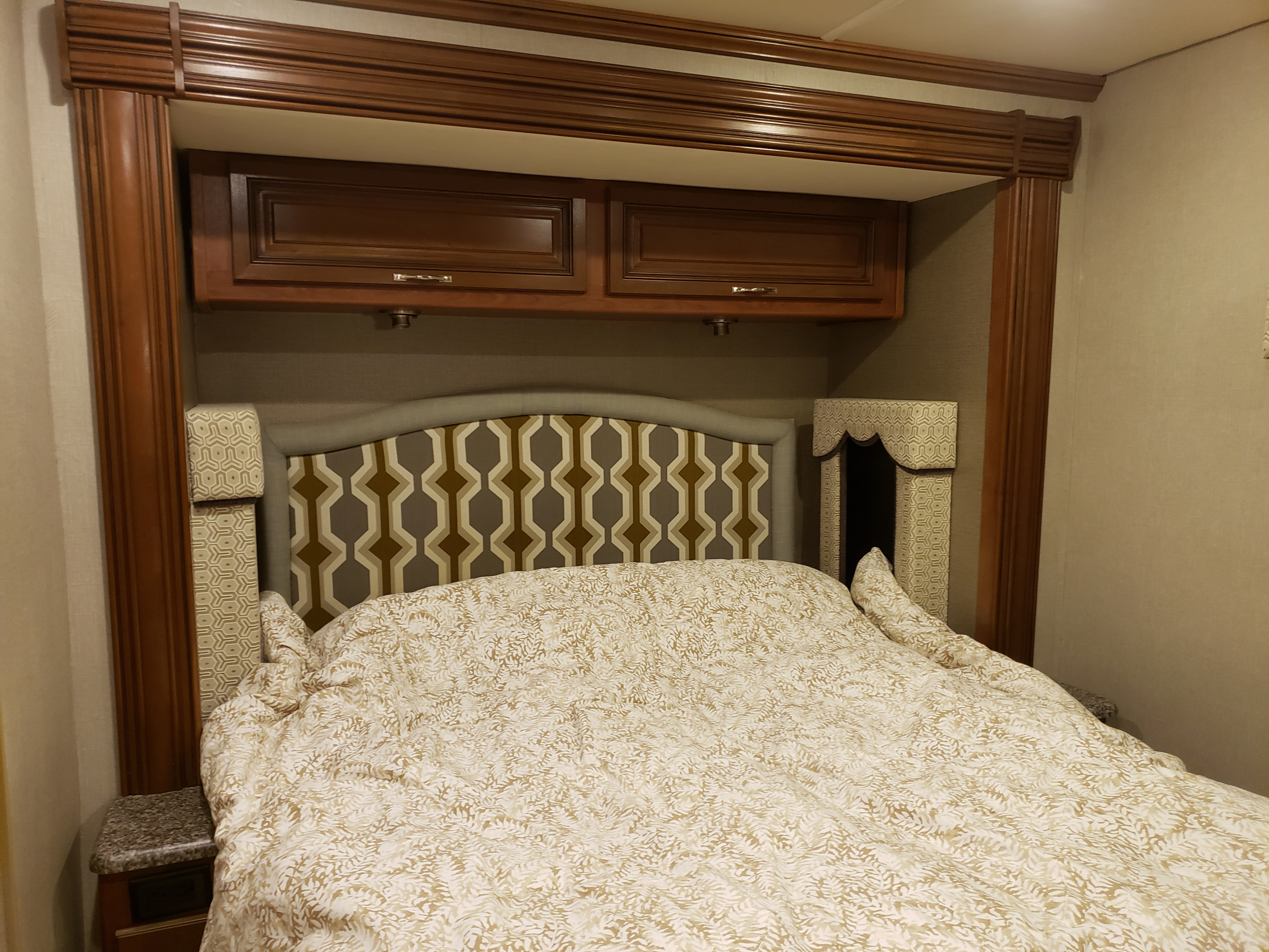 Cozy RV rental bedroom with a patterned headboard, light comforter, overhead wooden cabinets, and bedside storage.. Holiday Rambler Vacationer 2015