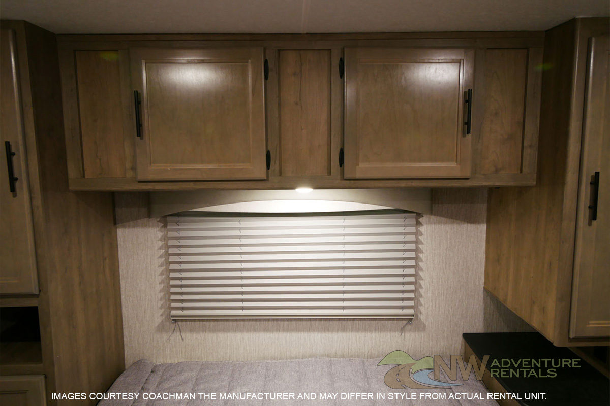 Cozy RV rental bedroom featuring wood overhead cabinets, under-cabinet lighting, a window with blinds, and a comfortable bed.. Coachmen Freelander 2020