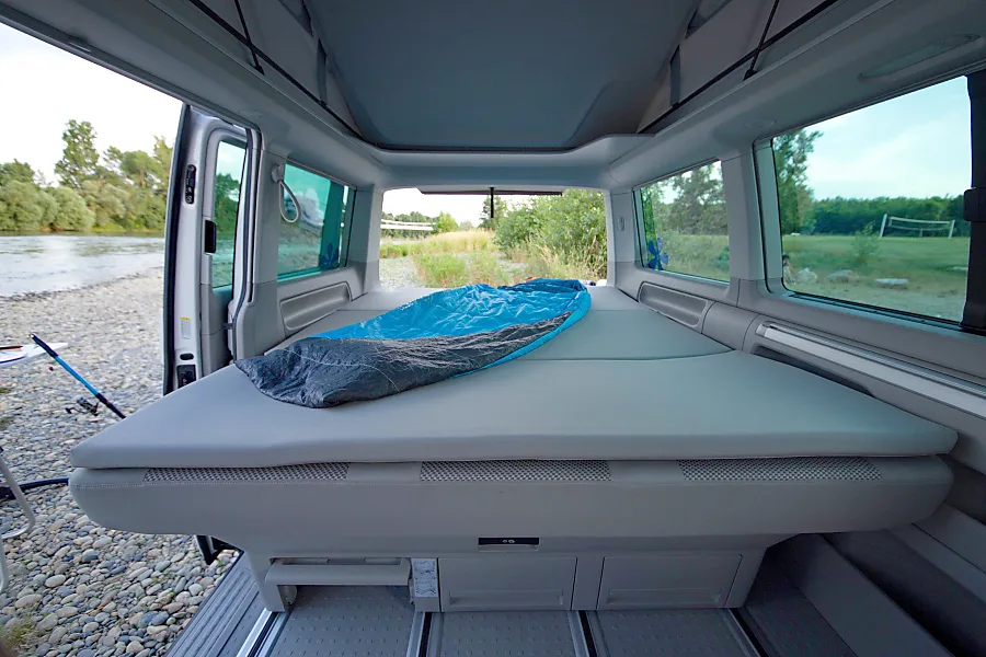 Inside a spacious RV rental: comfy bed with sleeping bag & serene riverside views through open doors.. Volkswagen California Beach T6.1 2023