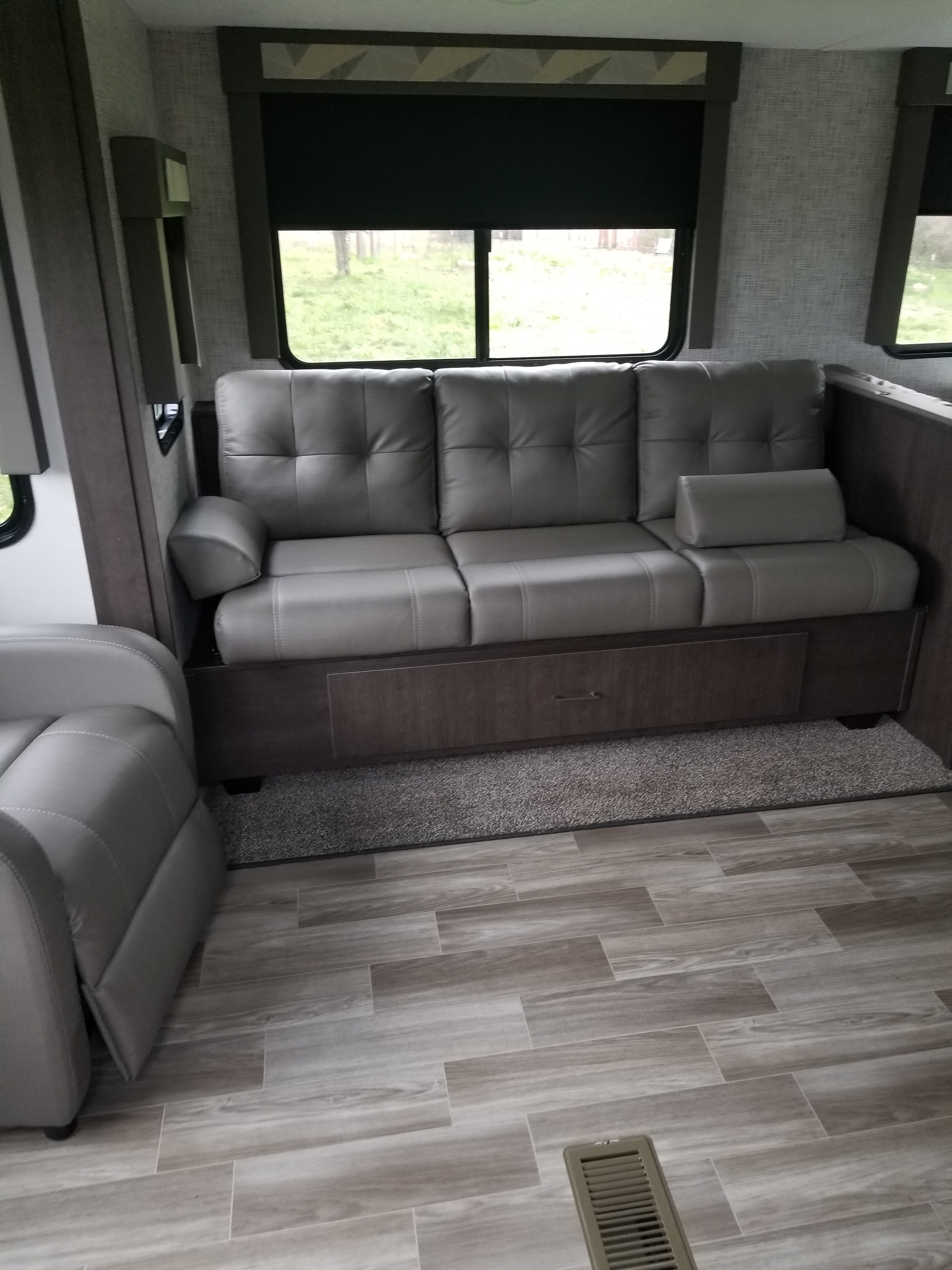 Comfy grey couch with storage in an RV rental. Wood-look floor, windows with blinds show outdoor greenery.. Forest River Wildwood 2019