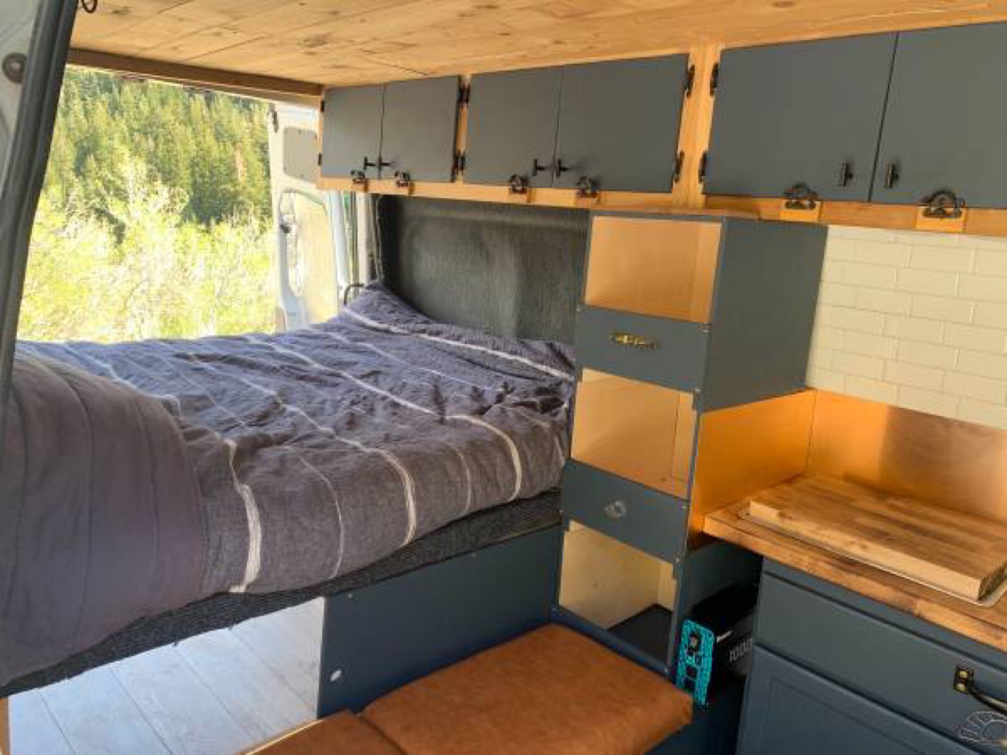 Cozy RV rental showing bed, overhead cabinets, kitchenette, and shelves. Outside, green trees suggest a nature escape.. Dodge Sprinter Van 2008