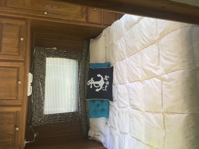 An RV rental sleeping area with a large white comforter, anchor pillow, window with blinds, and wood cabinets.. Ford E350 Box Van 2000