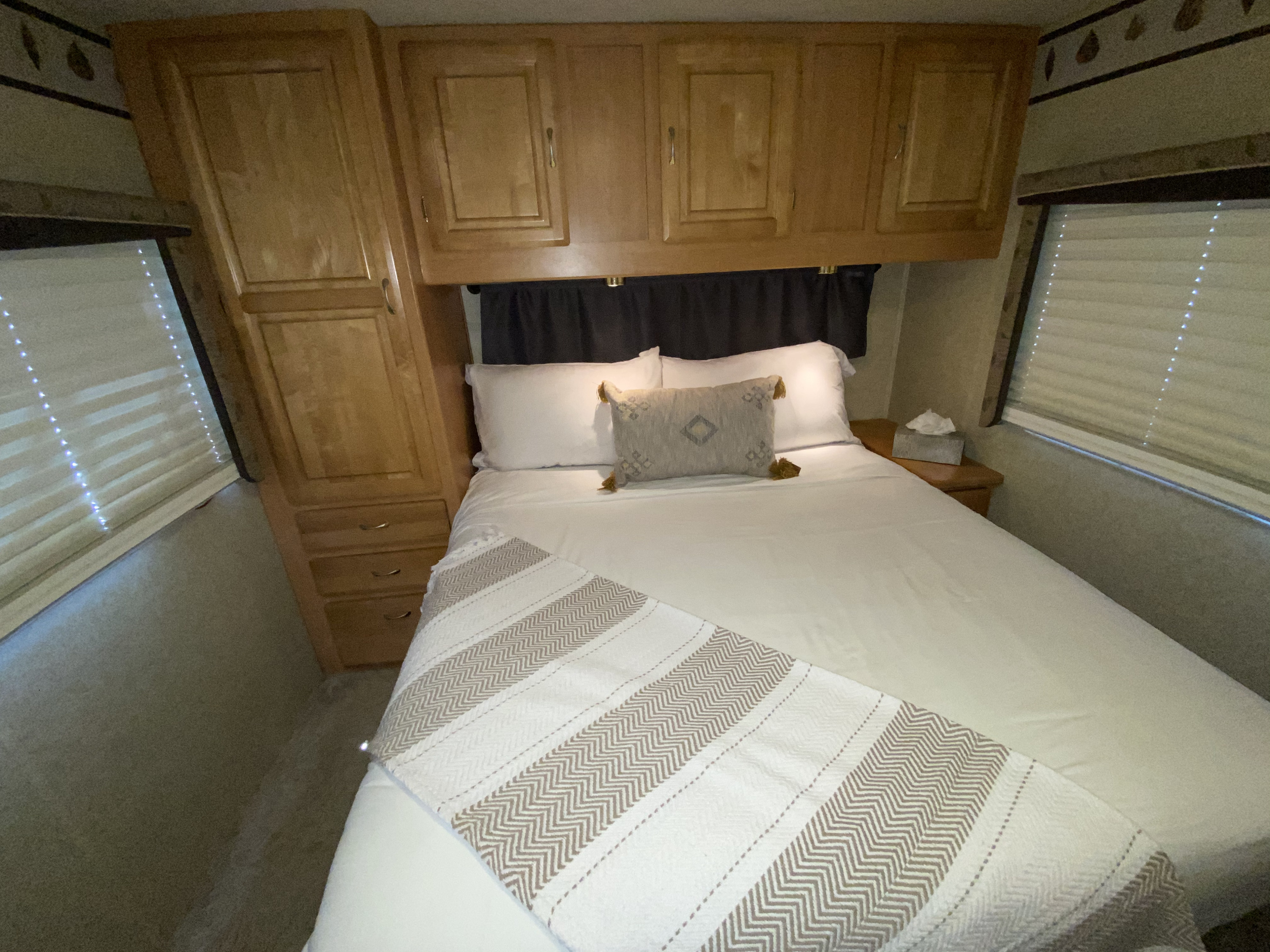 Cozy RV rental bedroom with queen bed, white linens, decorative throw, overhead cabinets, and window blinds. Ready for your adventure.. Jayco Granite Ridge 2003