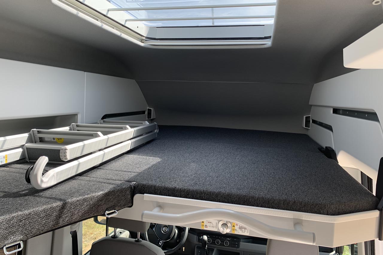 Overhead bed in a modern RV rental, showing a dark grey mattress and a folded ladder for access.. Volkswagen Grand California 2021