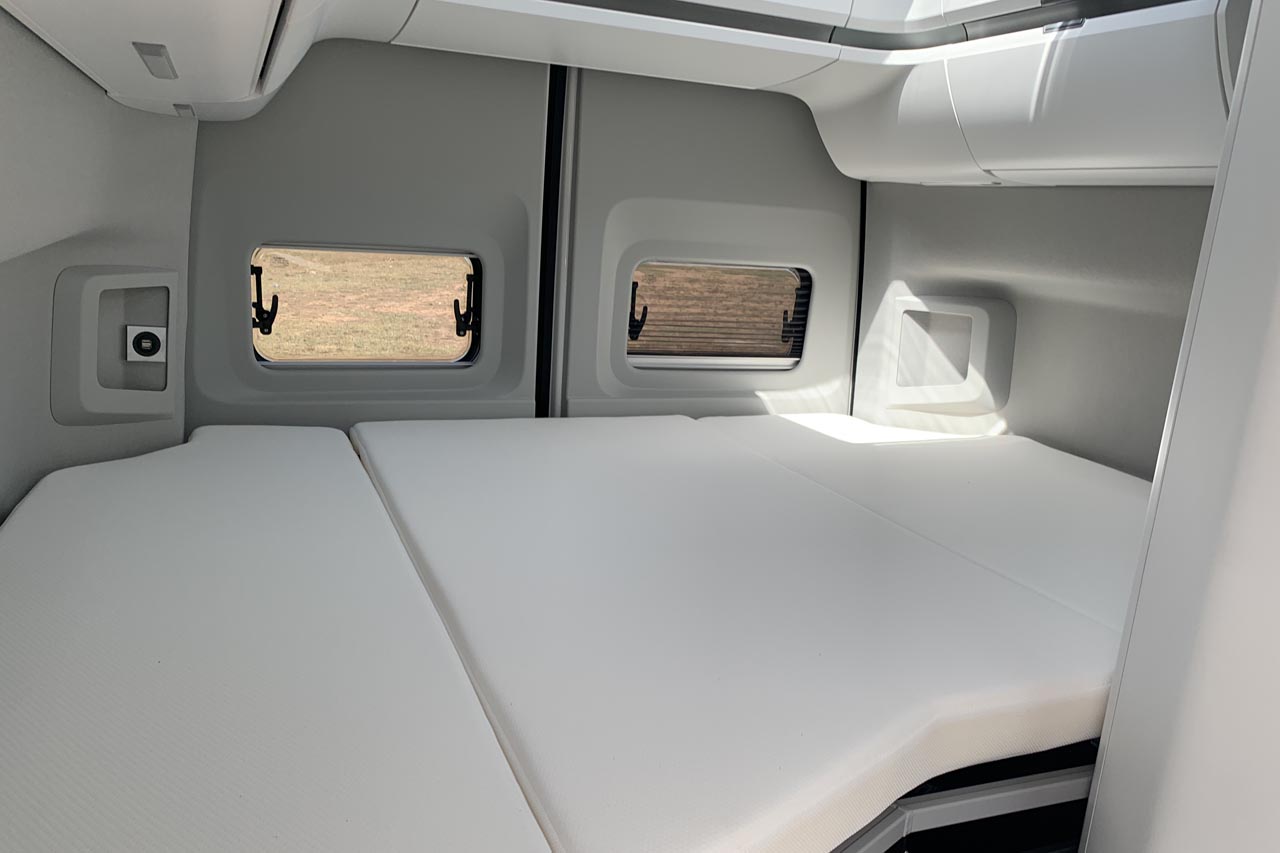 Inside a bright RV rental: a large, comfortable bed, side windows, and modern interior design for your travels.. Volkswagen Grand California 2021