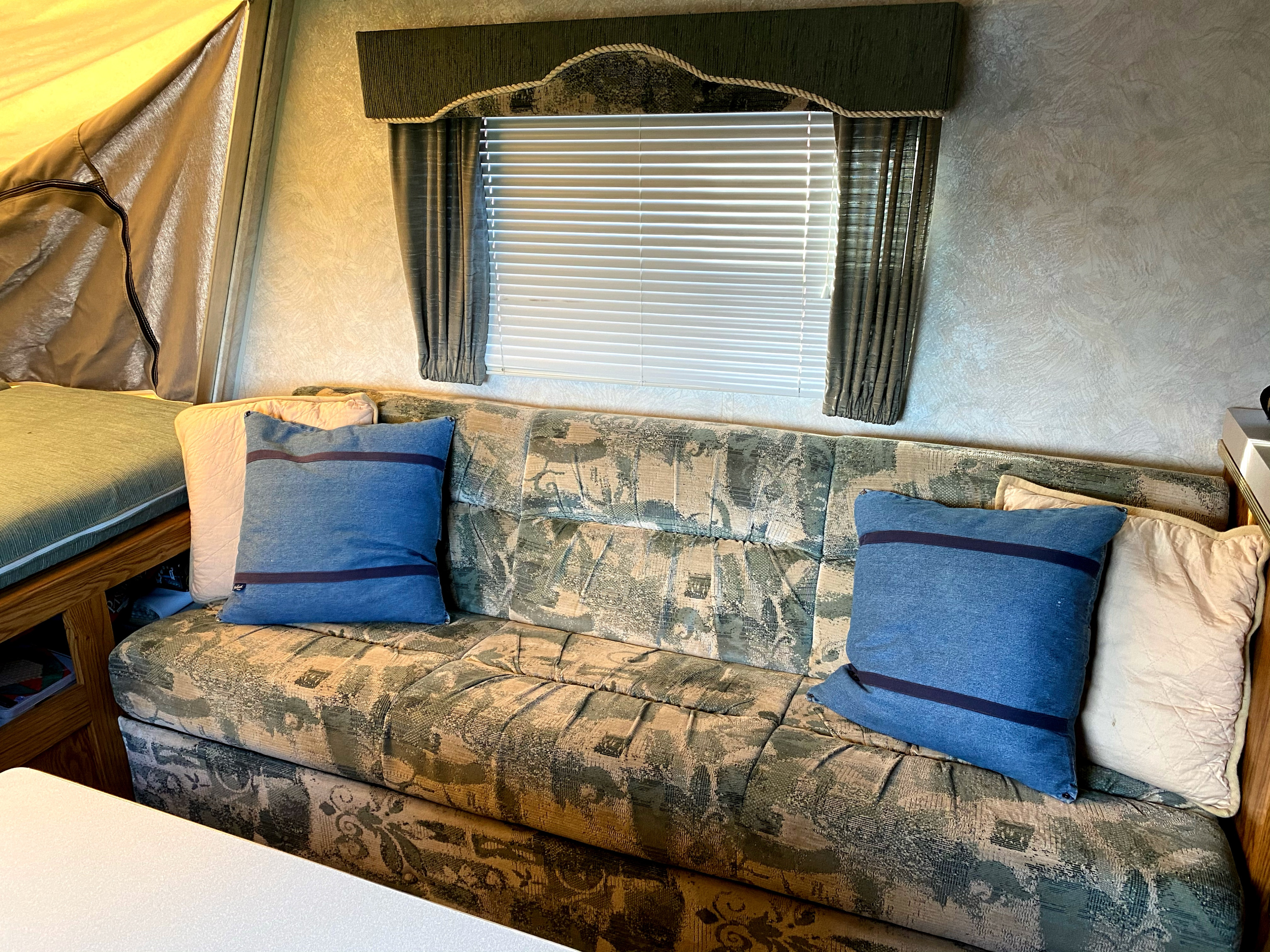 Cozy RV rental interior featuring a patterned sofa, blue pillows, window with blinds, and pop-up bed area.. Sportsmen Coyote 2001