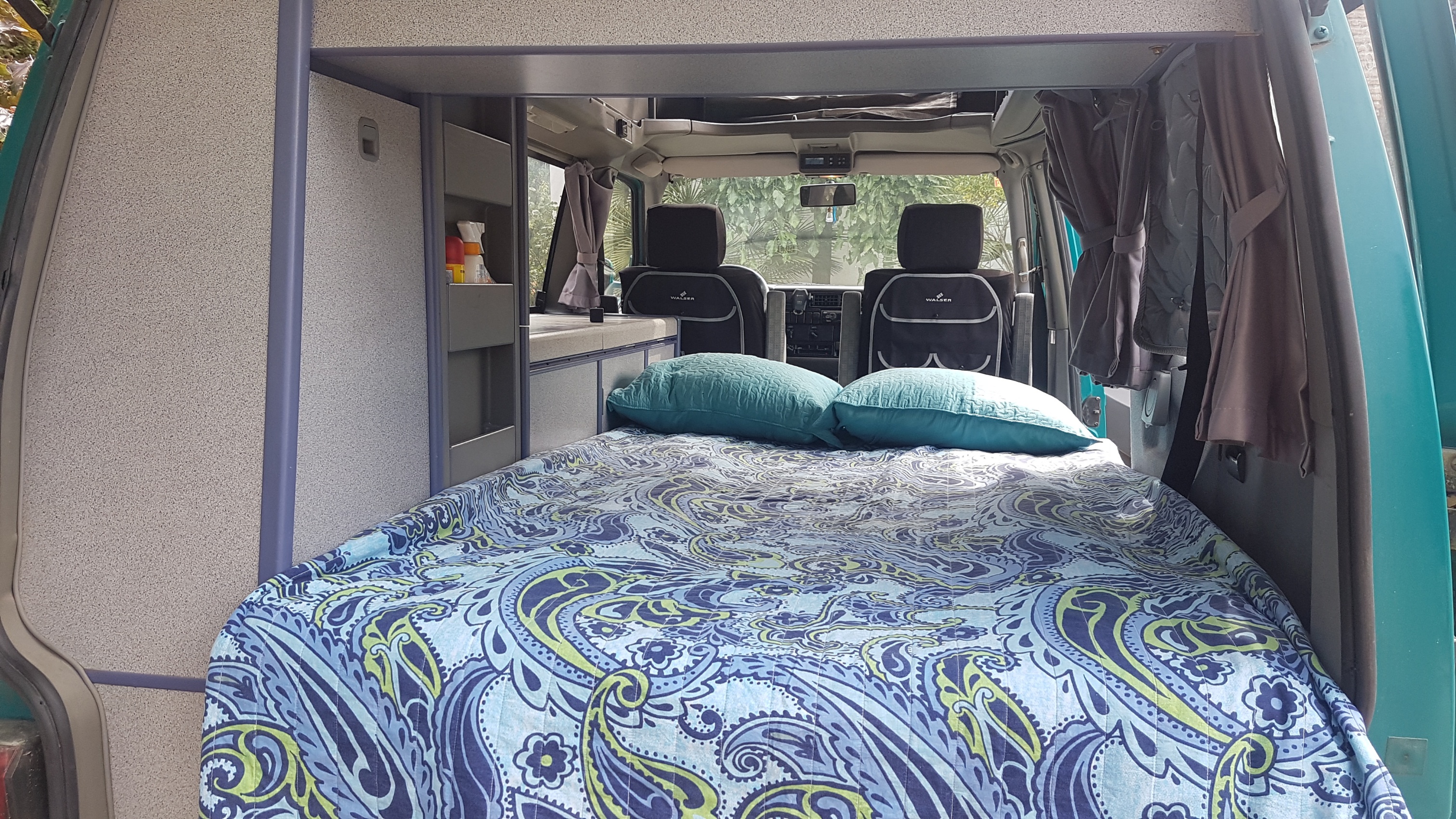 Cozy RV rental interior: a comfortable patterned bed with pillows, looking towards the driver's cabin and built-in storage.. Volkswagen California - Westfalia 1996