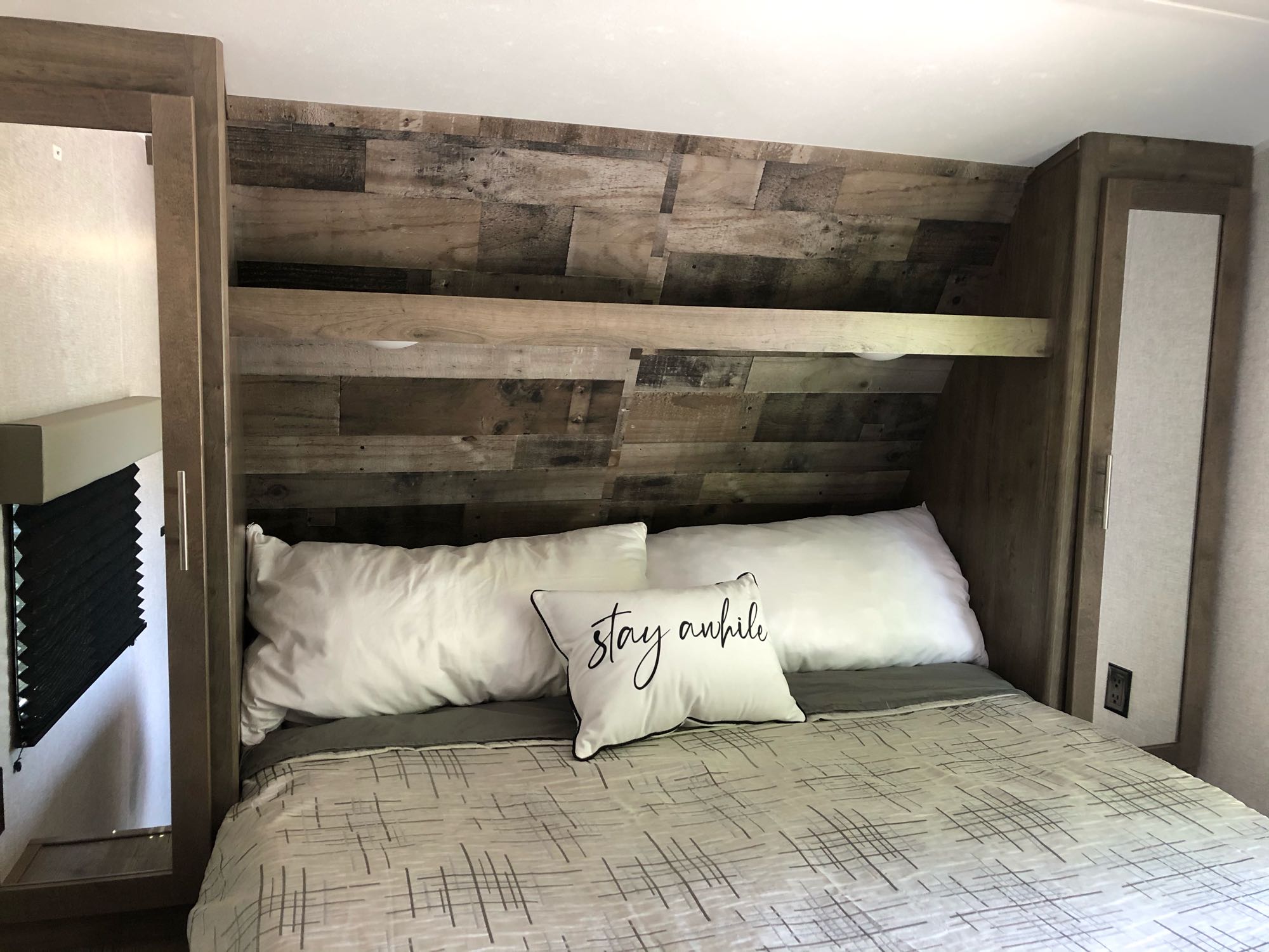 Cozy RV rental bedroom featuring a bed with white pillows, a "stay awhile" pillow, and rustic wood-paneled walls.. Dutchmen Aspen Trail 2020