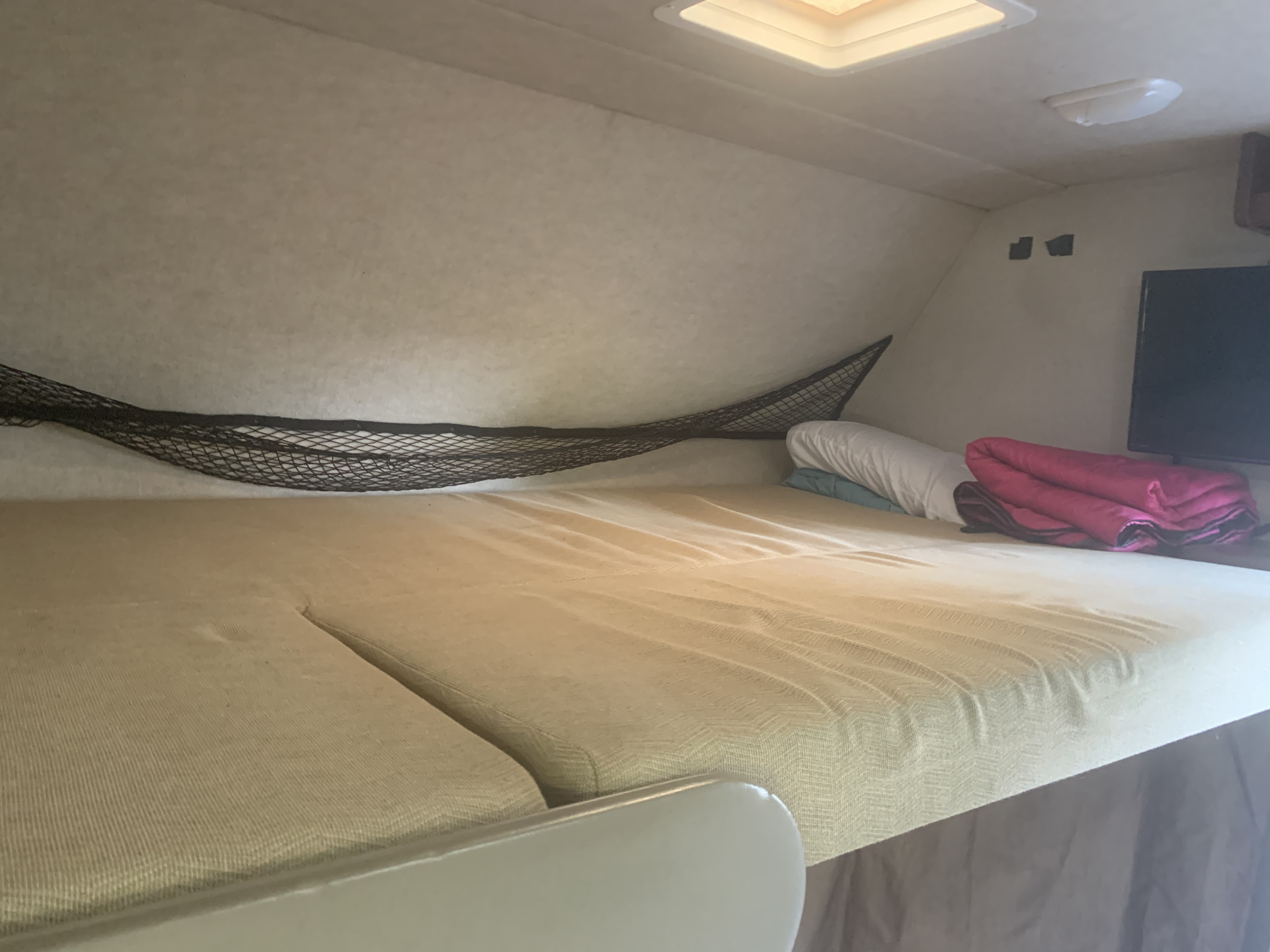 Inside an RV rental, a neatly made overhead bunk bed features light bedding, a safety net, pillows, and a TV for relaxation.. Fleetwood Jamboree Searcher 2013