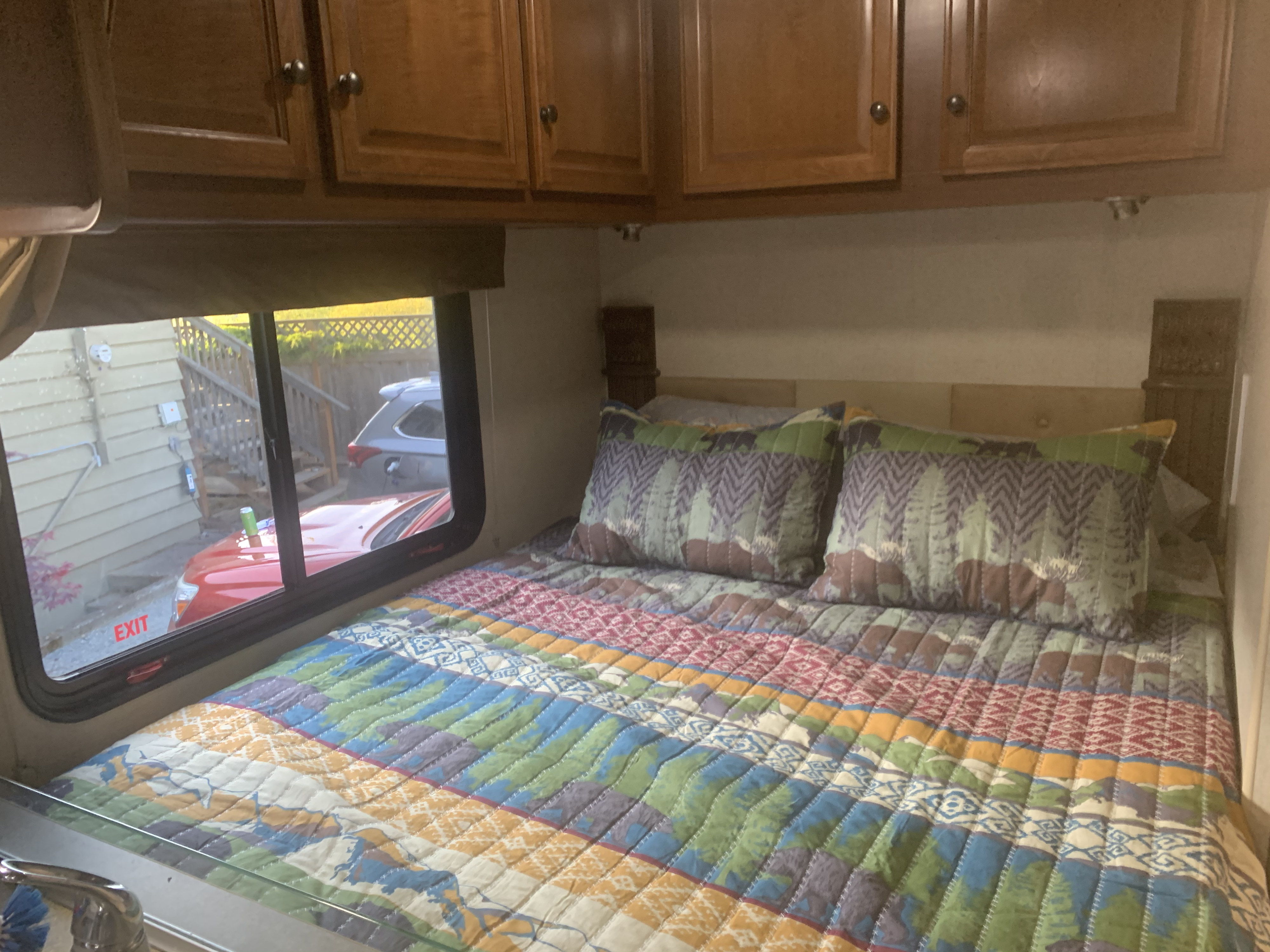 Cozy RV rental bedroom with a vibrant, patterned quilt, pillows, and overhead cabinets. Window view includes an EXIT sign.. Fleetwood Jamboree Searcher 2013