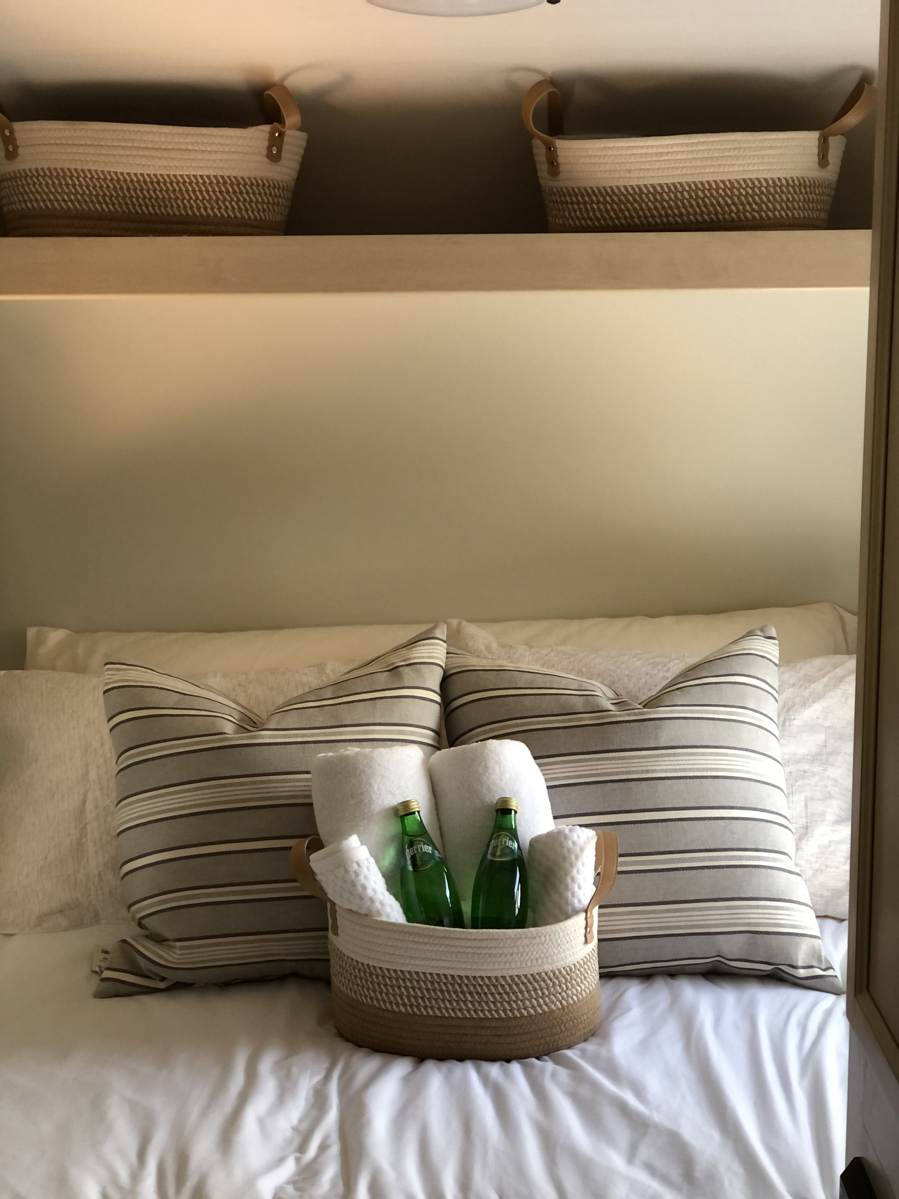 Cozy RV rental bed setup with striped pillows, welcome basket, rolled towels, and Perrier water bottles.. Winnebago Minnie Winnie 2018