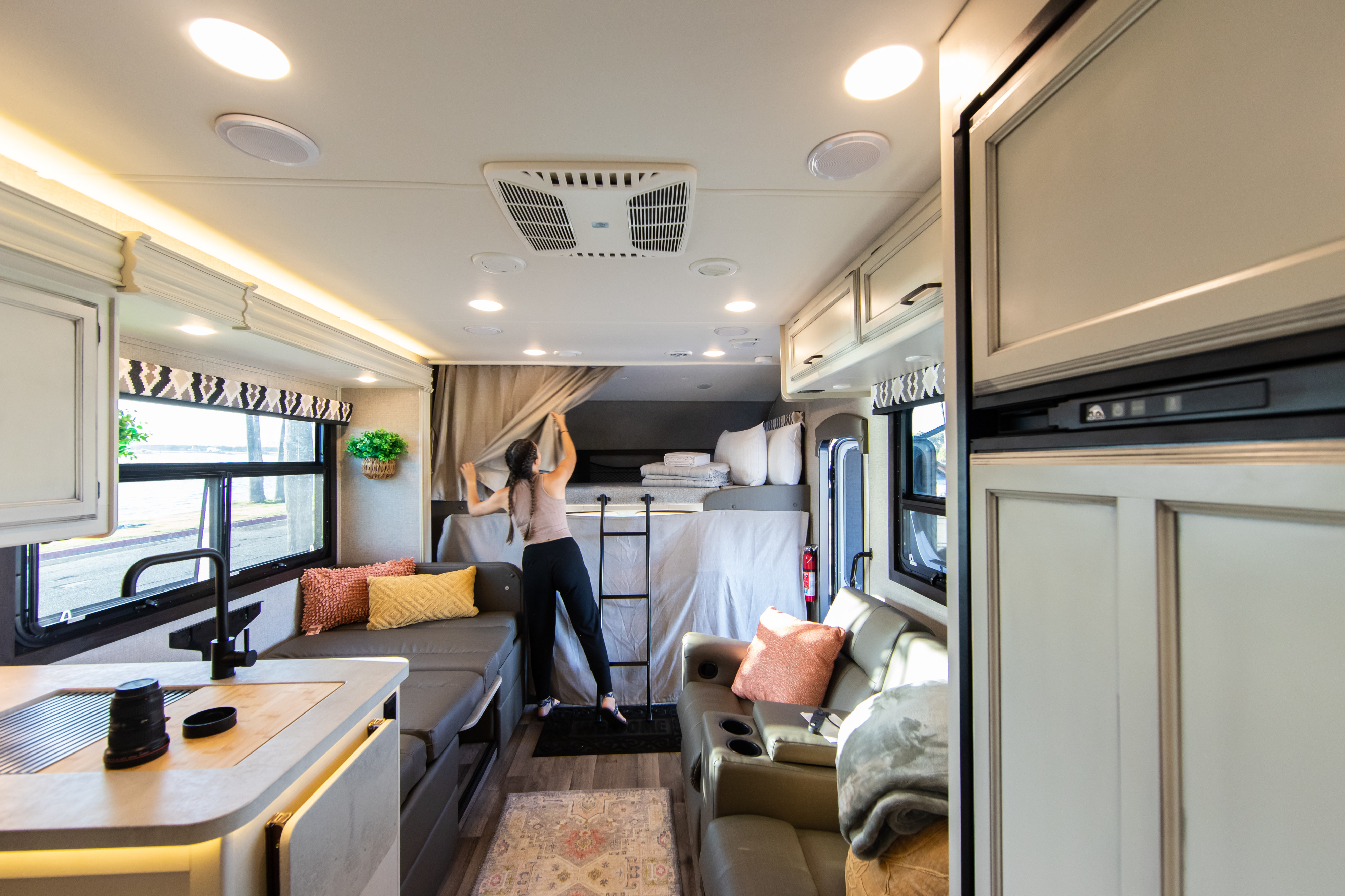 Modern RV rental interior: A person arranges curtains by the bunk bed, showcasing the kitchen, lounge, and amenities.. Jayco Greyhawk 2021