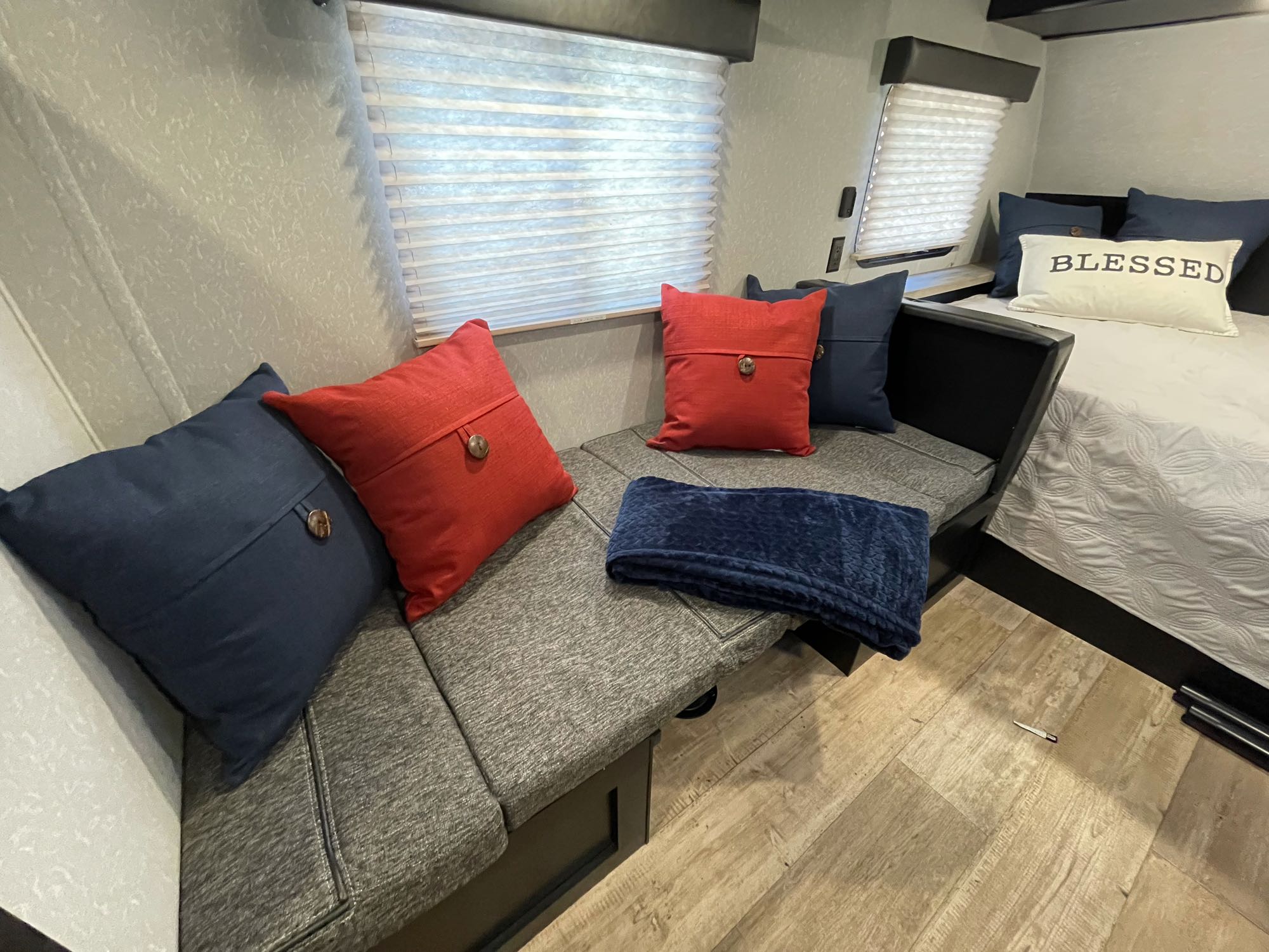 Cozy RV rental interior shows a grey L-shaped dinette with red and navy pillows, a blue blanket, and a bed with a "BLESSED" pillow. Ready for travel.. Forest River Wolf Den 16EV 2021
