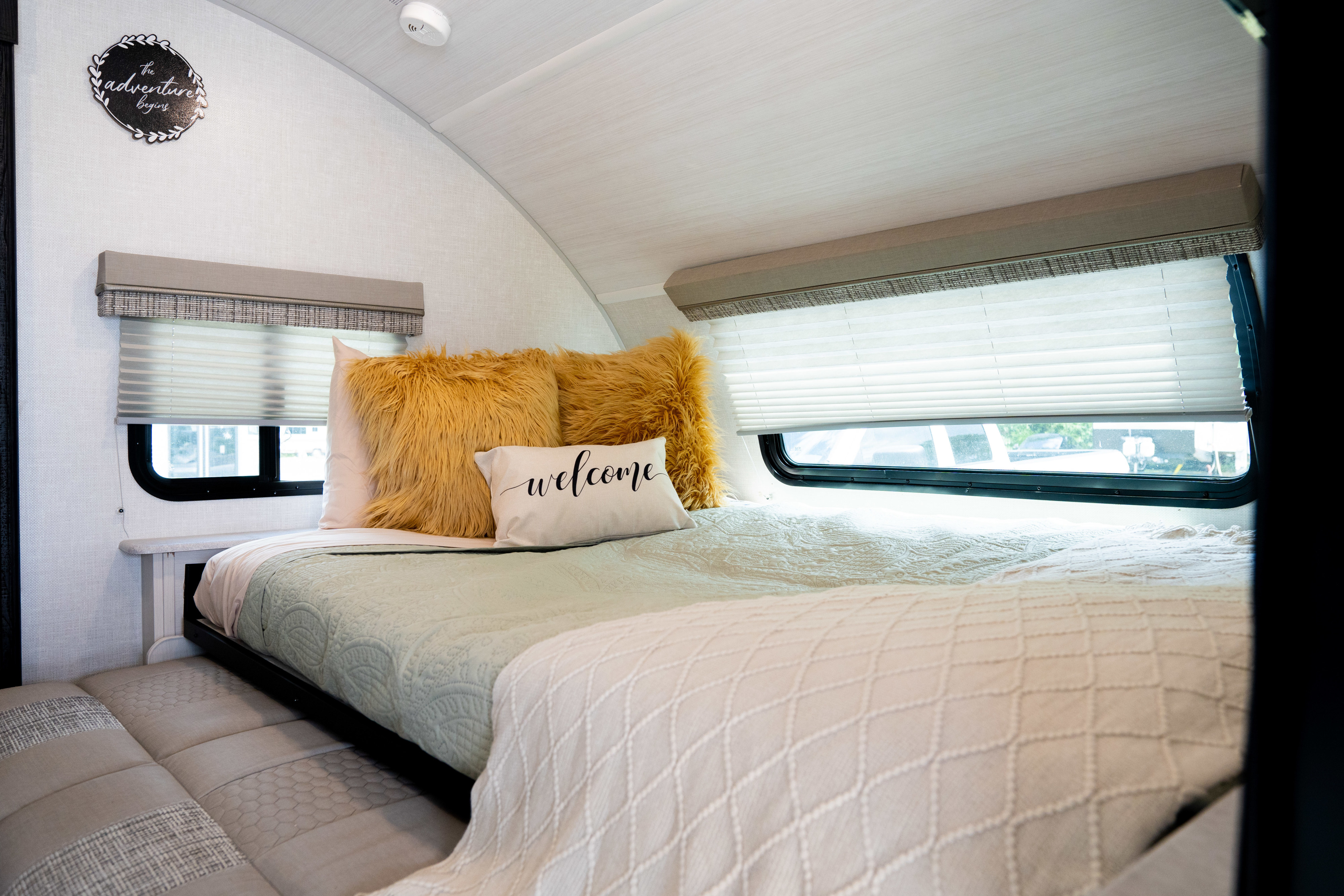 Cozy RV rental bedroom with light green bedding, fluffy yellow pillows, a "welcome" pillow, windows, and "adventure" sign.. Forest River R-Pod 2021