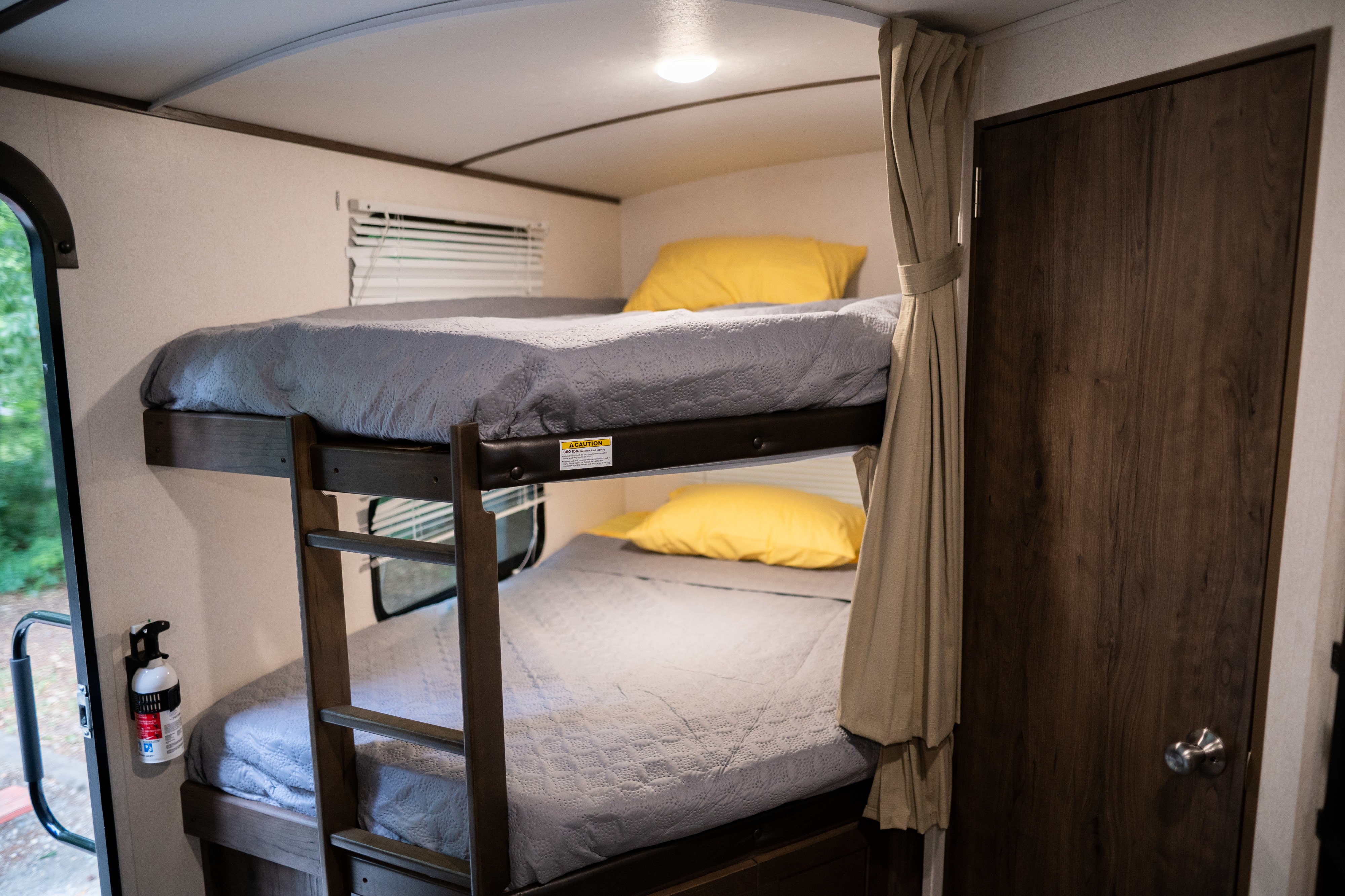 Interior view of an RV rental showing stacked bunk beds with grey quilts & bright yellow pillows, plus a fire extinguisher.. Keystone Passport 2020