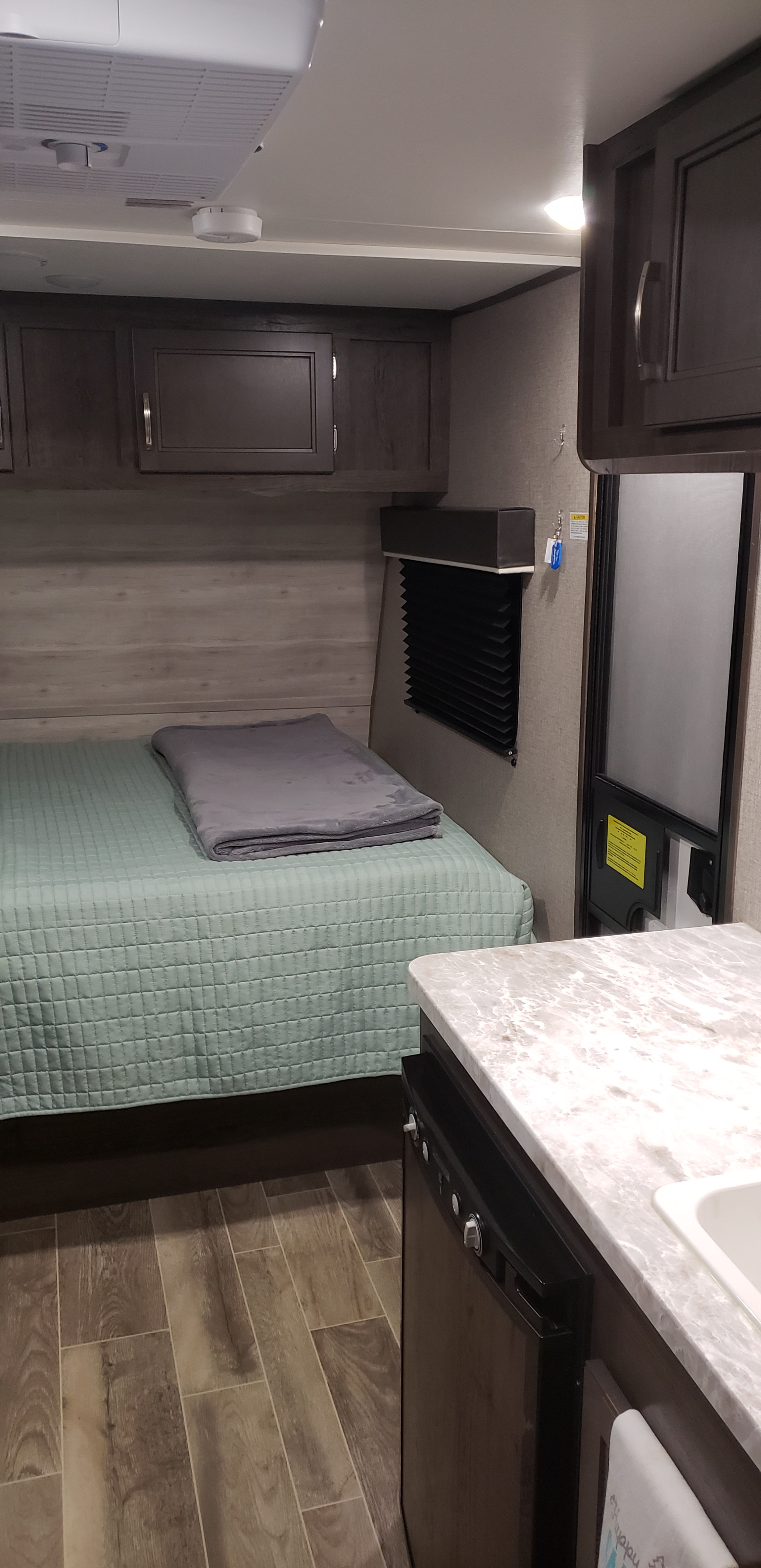 Inside an RV rental: a comfy bed, wood-look floor, overhead cabinets, and a compact kitchen counter with a fridge.. Jayco Jay Flight 2021