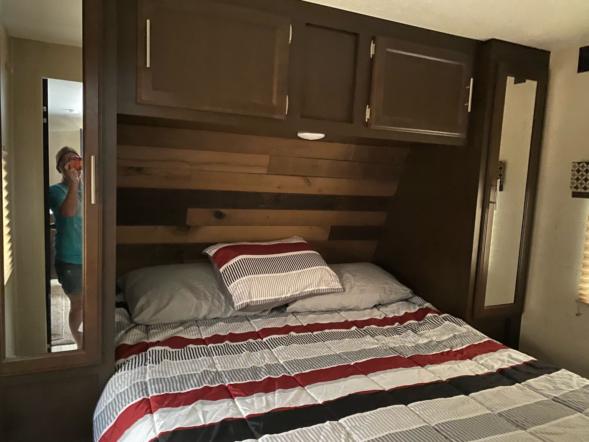 Cozy RV rental bedroom with a striped bed, overhead cabinets, and built-in mirrors reflecting a person.. K-Z Manufacturing Sportsmen 2018