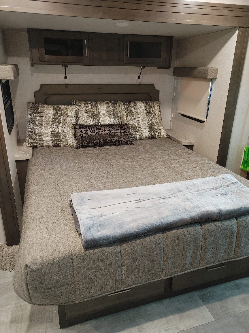 Cozy RV rental bedroom showing a queen bed with patterned pillows, overhead cabinets, and soft lighting. Ideal for travel comfort.. Forest River Rockwood Ultra Lite 2021