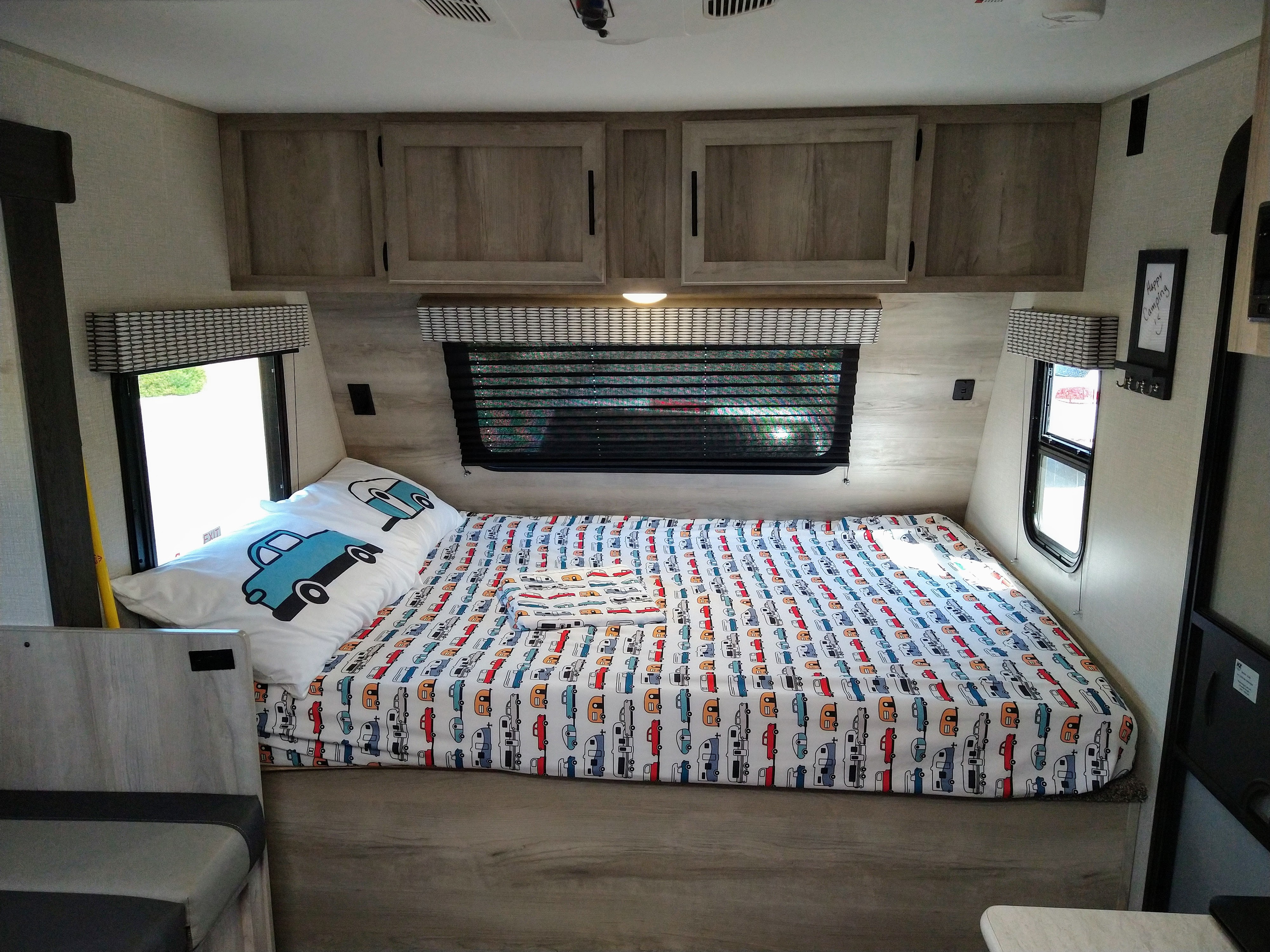 Cozy RV rental bed with blue truck pillow & playful camper-patterned sheets. Bright, functional interior.. KZ 181BH 2021
