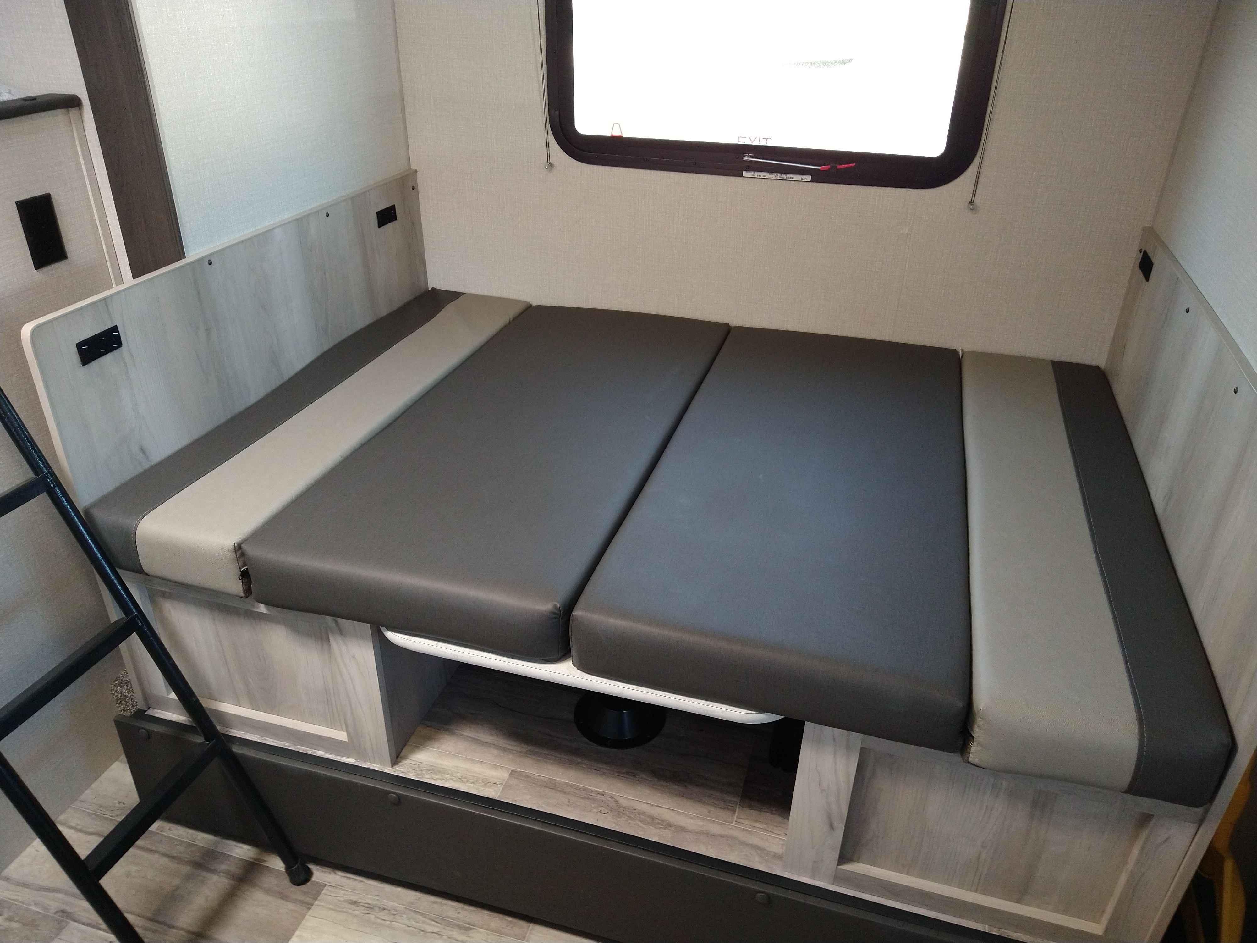 An RV rental interior showing a versatile bunk bed/dinette with grey cushions, built-in storage, and an emergency exit window.. KZ 181BH 2021