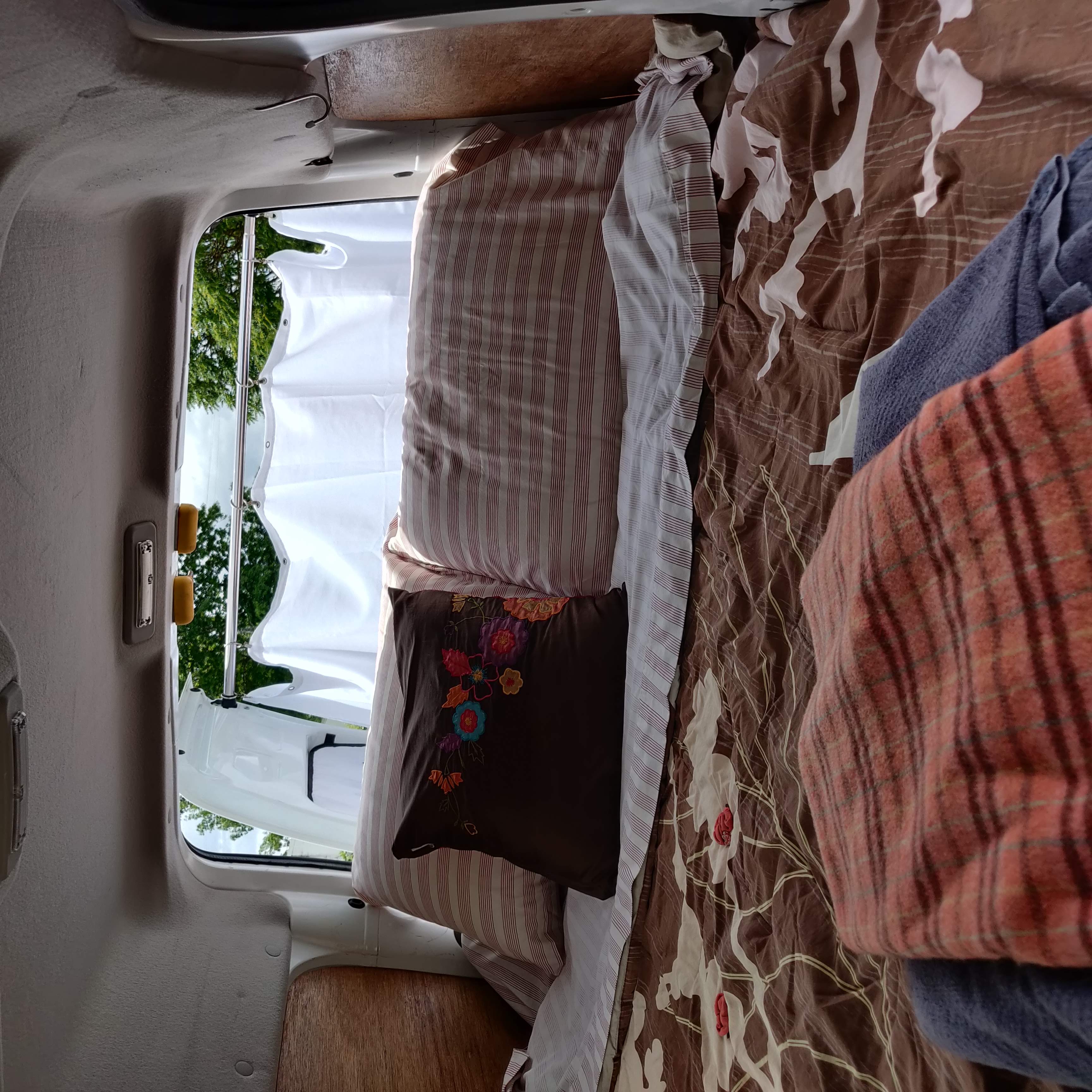 Inside a cozy RV rental: a comfortable bed with colorful pillows, offering a scenic view out the window.. Ford Connect 2012