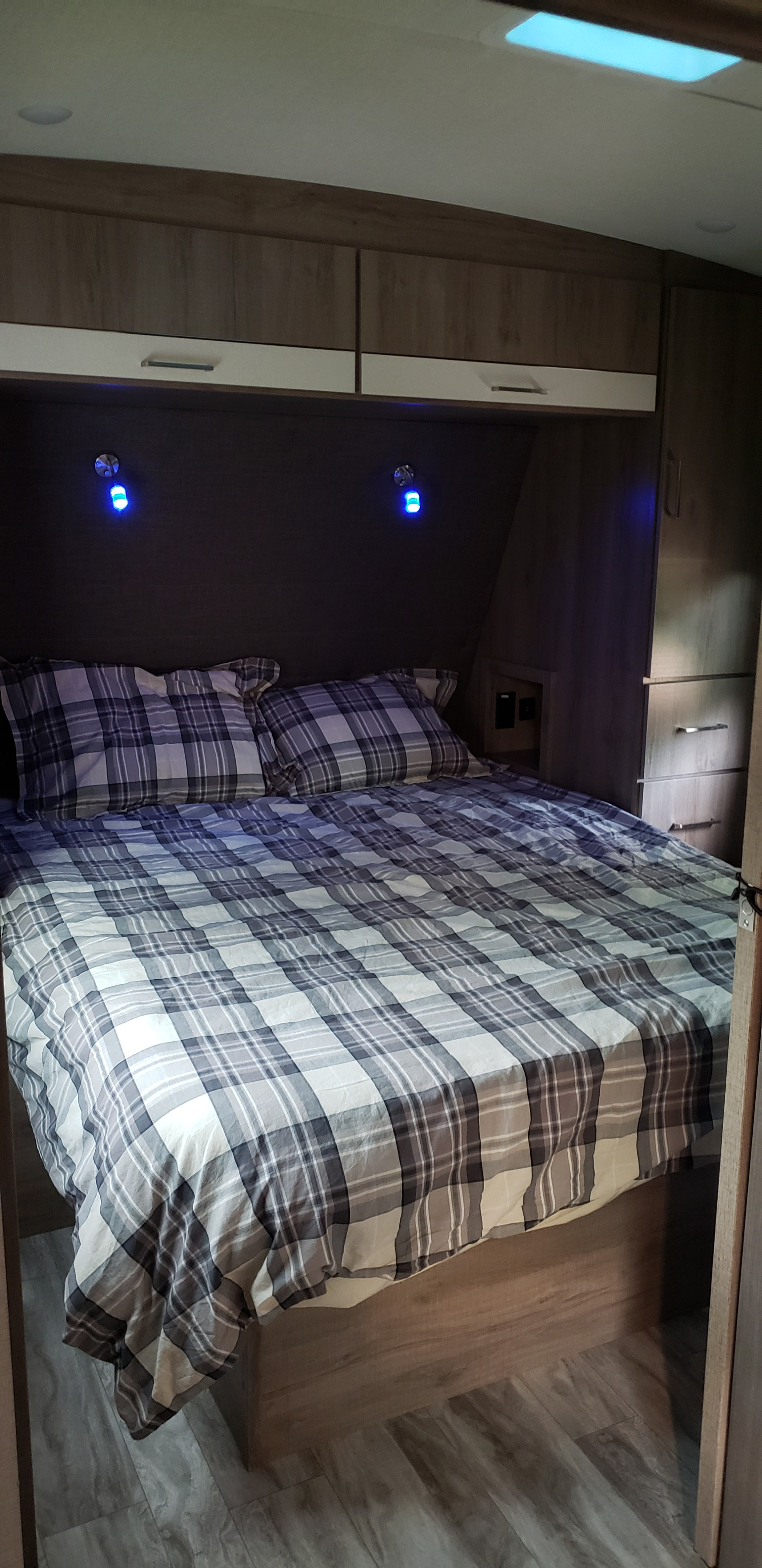 A modern RV rental bedroom shows a plaid bed, built-in overhead cabinets, side dresser, and blue accent lights for a compact interior.. Grand Design Imagine 2400BH 2021
