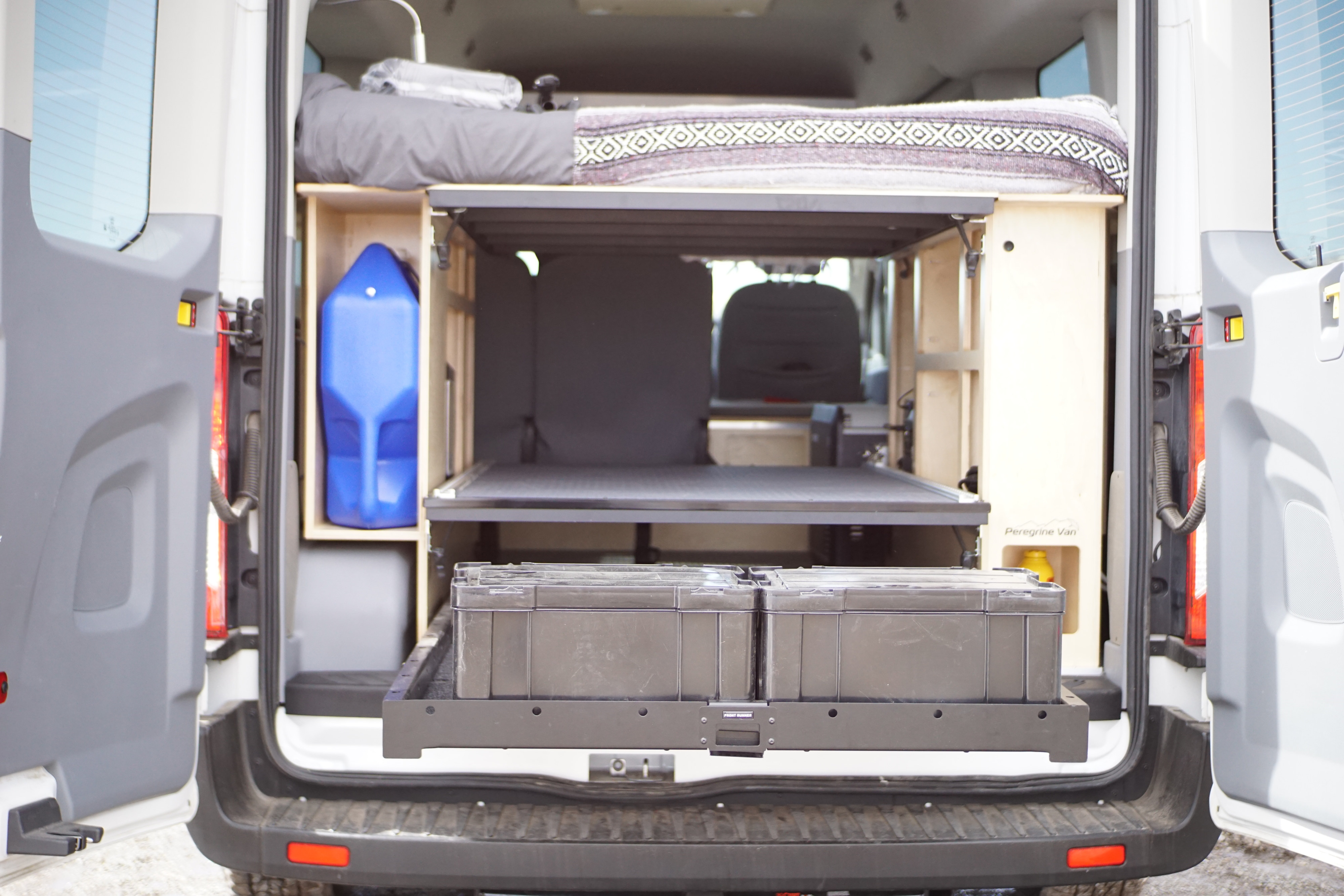 Inside this rv rental: a camper van interior with an elevated bed, pull-out storage crates, and a blue water jug for road trips.. Ford Transit Custom 2019