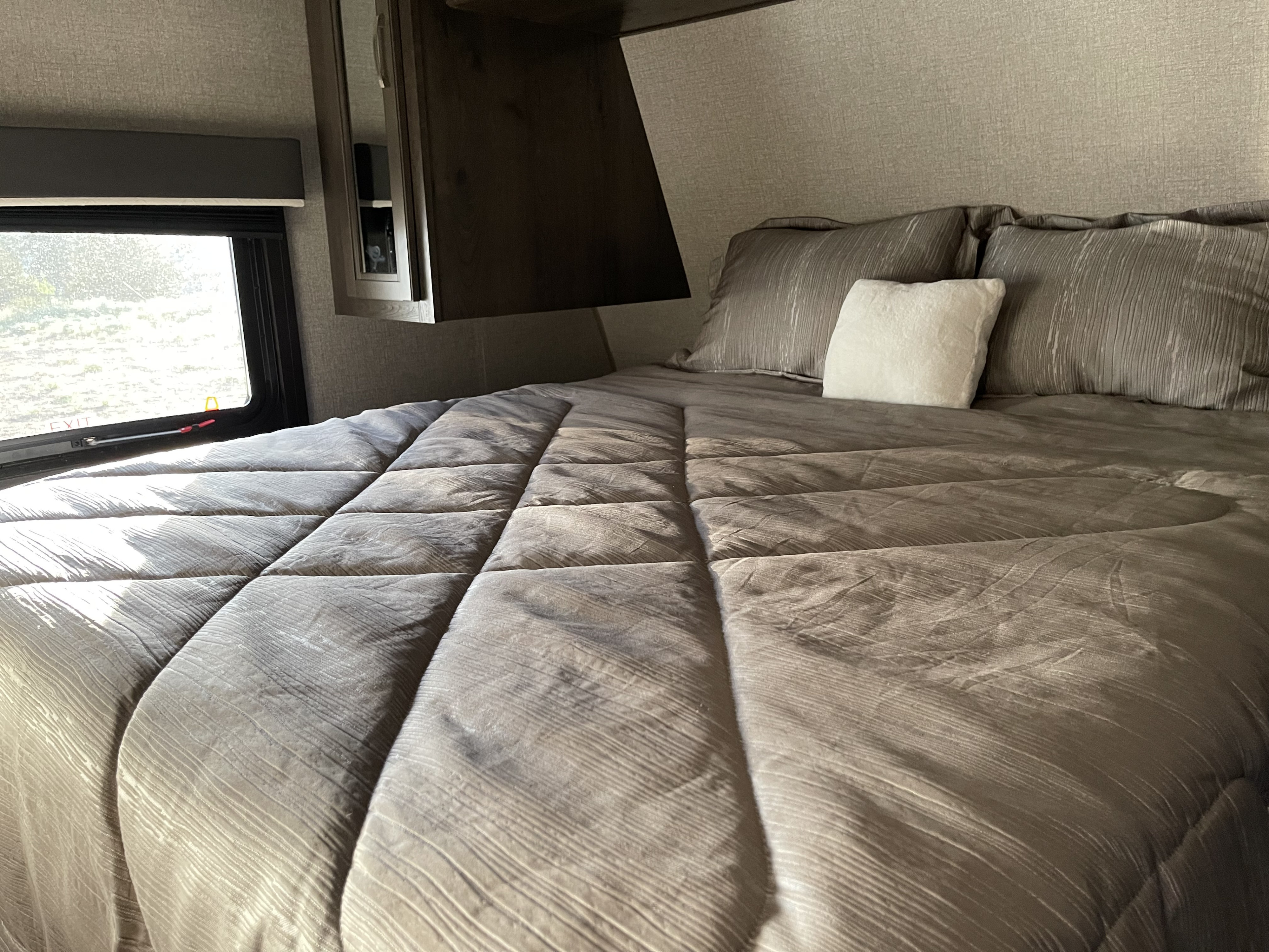Experience comfort in this RV rental bedroom: plush gray bedding, a large window with sunlight, and overhead cabinets.. Jayco Jay Flight 2020