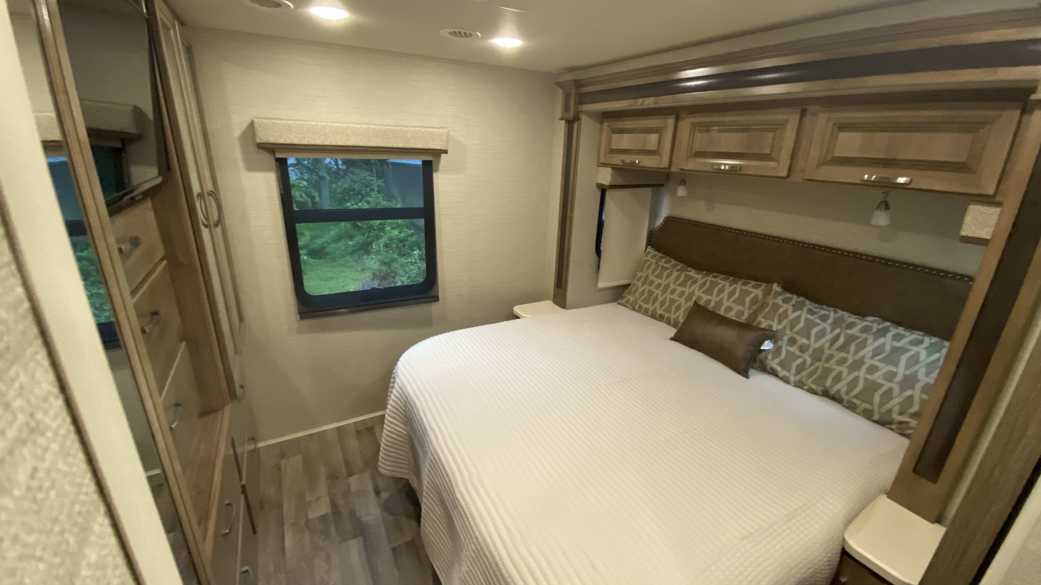 This inviting RV rental bedroom features a cozy bed with pillows, overhead cabinets, a window with an outdoor view, and wardrobe storage.. Jayco Precept 2019