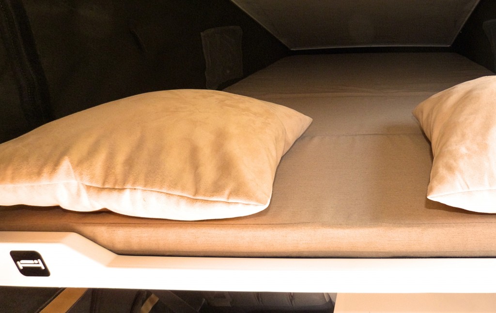Clean and comfy bed setup inside an RV rental. A mattress and two light-colored pillows provide a cozy sleeping space.. Jeep Rubicon Pop-top 2016
