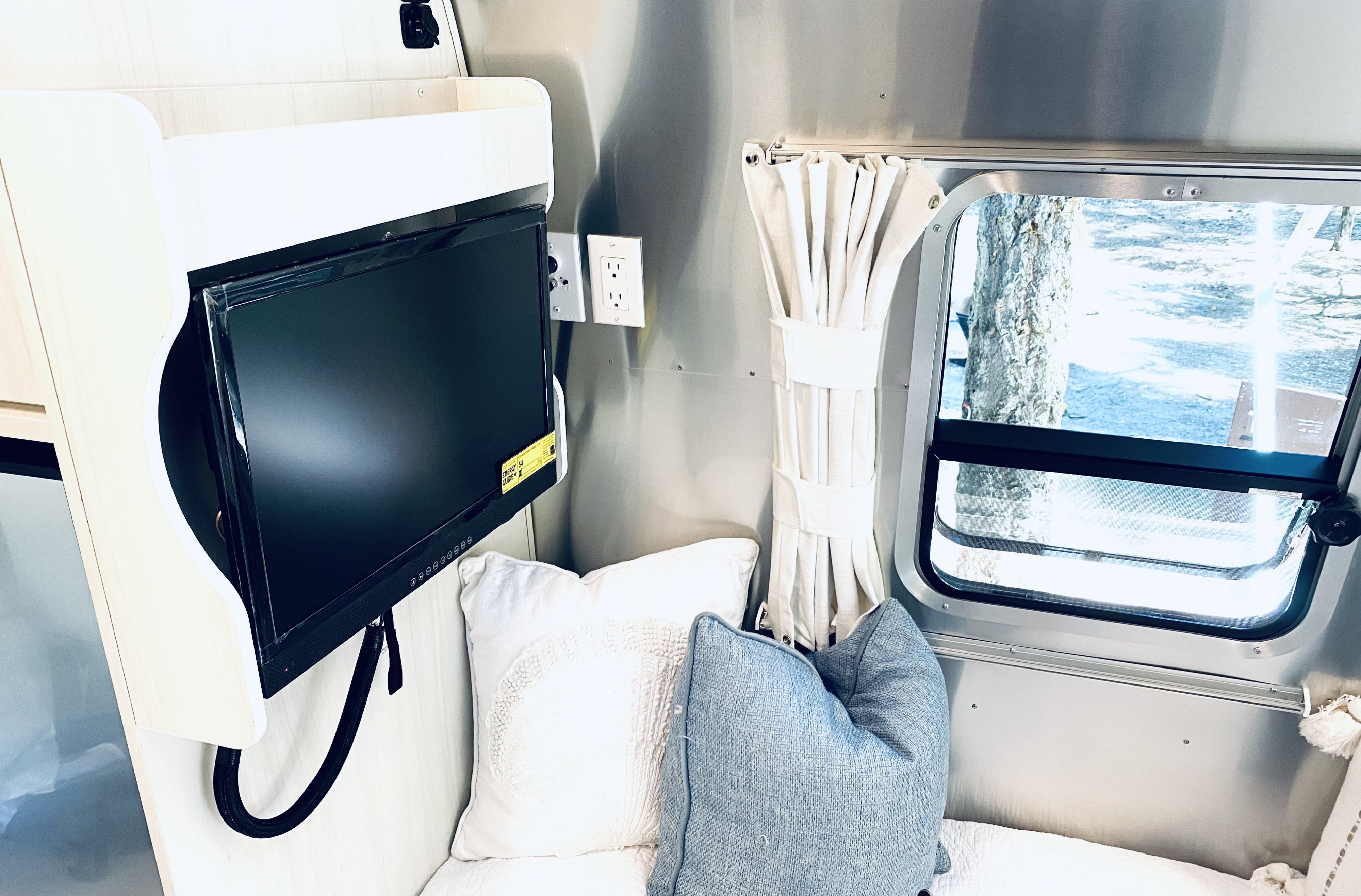 Cozy RV rental interior featuring a TV, plush pillows, and a large window showing a snowy treeline outside.. Airstream Bambi 2022