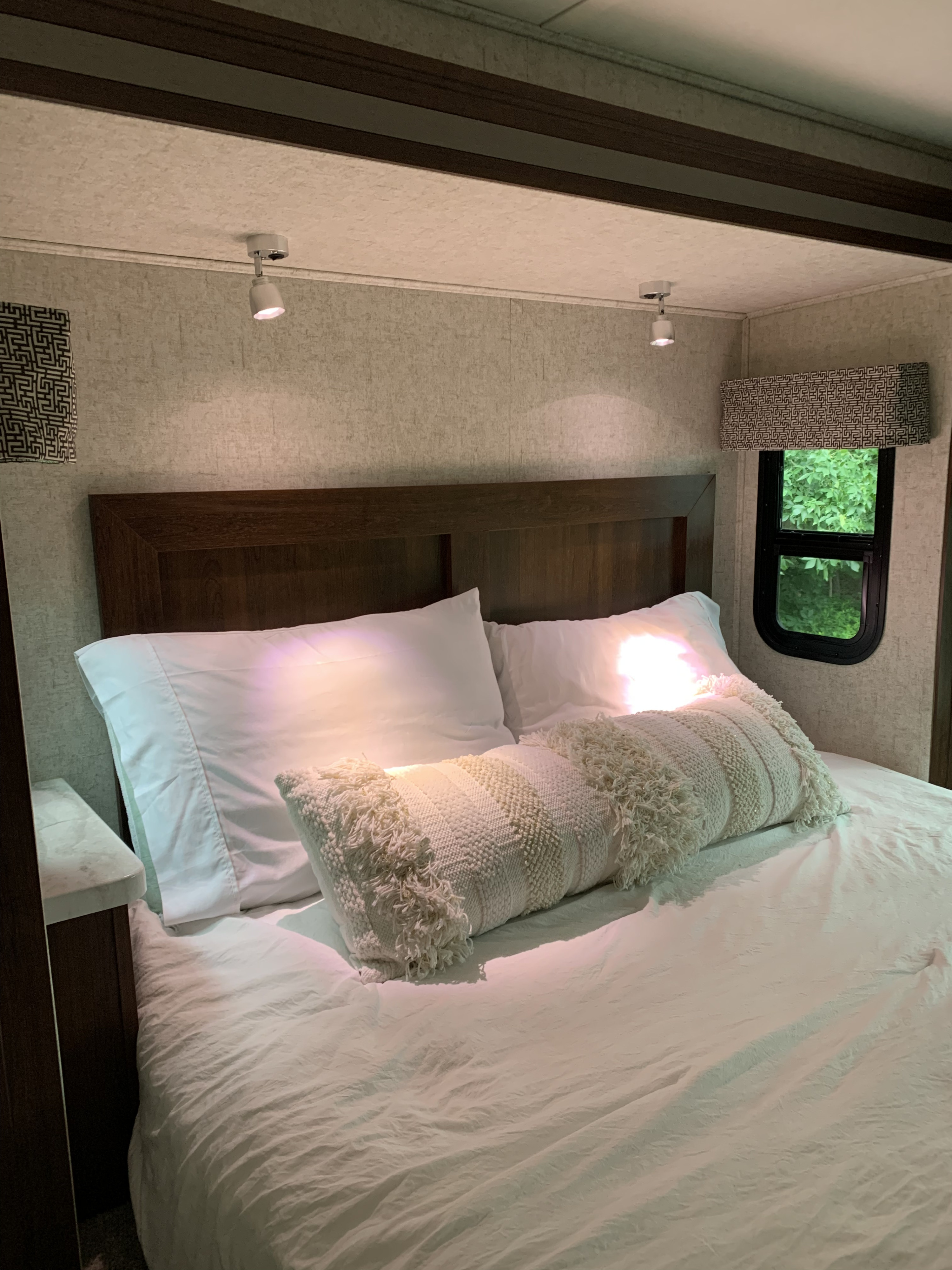 Cozy RV rental bedroom featuring a bed with white linens, a decorative long pillow, spotlights, and a window with a leafy view.. Forest River Sunseeker 2018