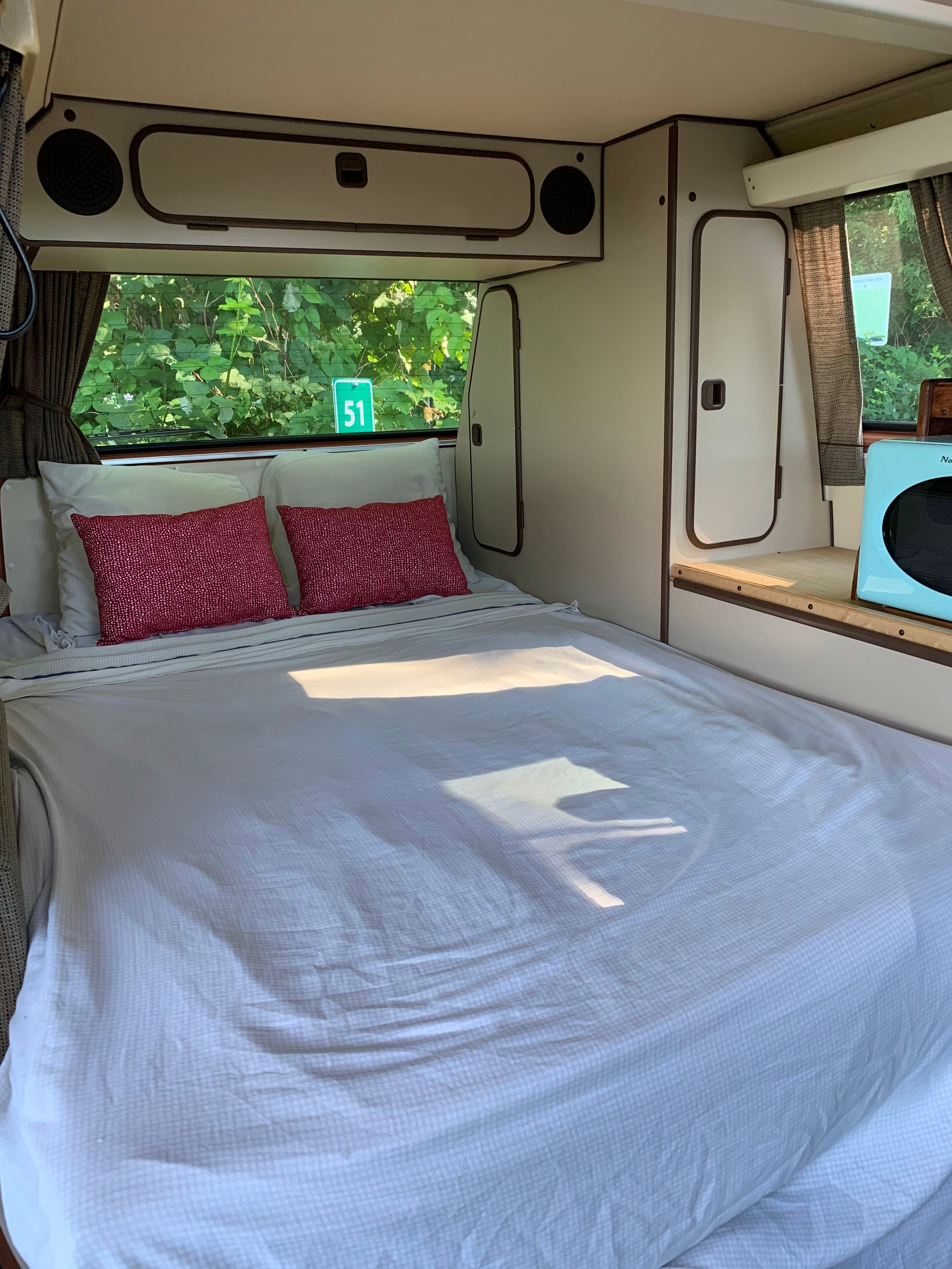 Cozy RV rental bedroom with a comfortable bed, red pillows, overhead storage, and small kitchen area with a microwave.. Volkswagen Vanagon 1984
