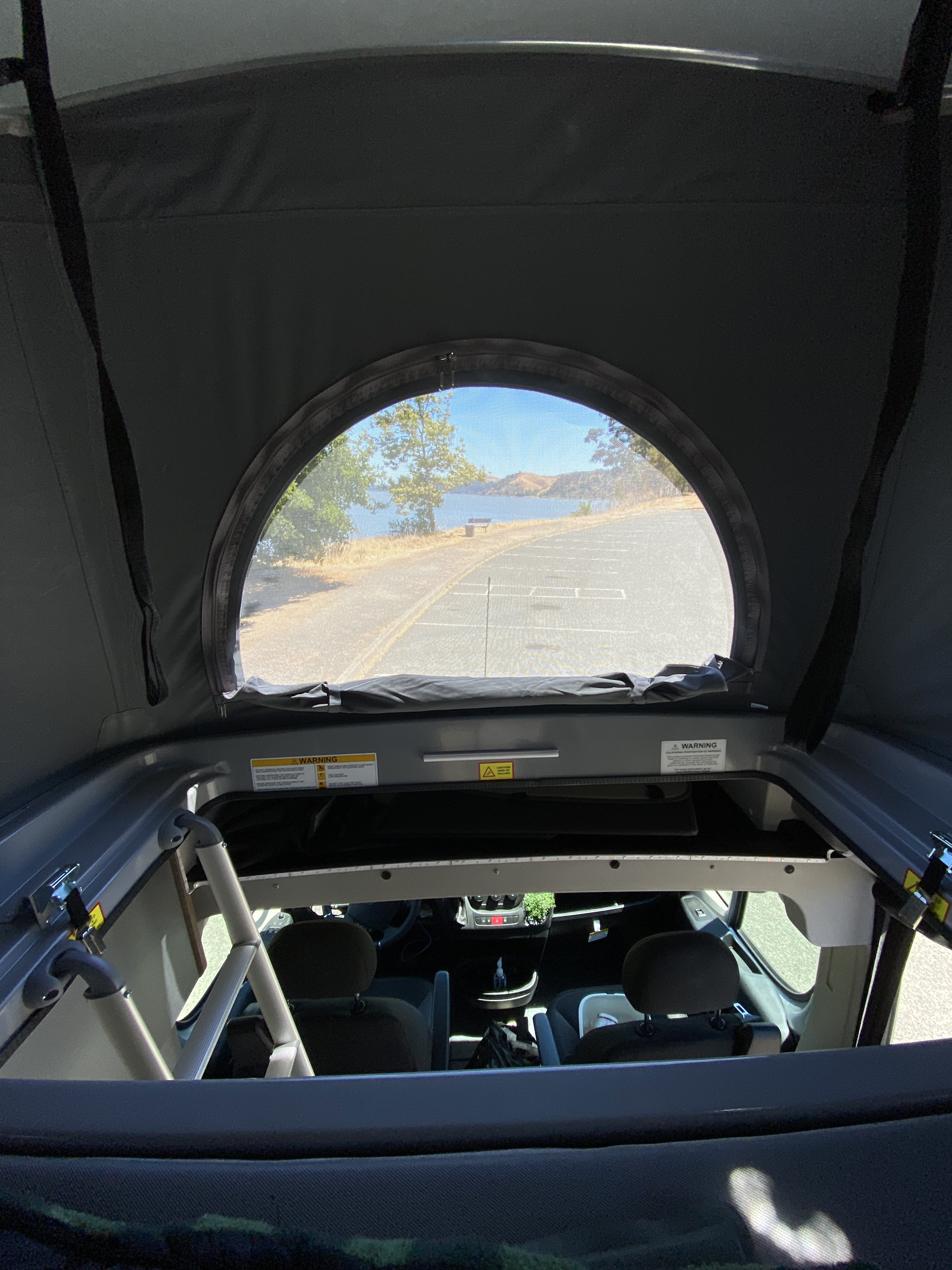 RV rental interior: View from a pop-top bed area, looking through a screen window at a scenic lake, road, & distant hills. The RV's cab is visible below.. Winnebago Other 2022