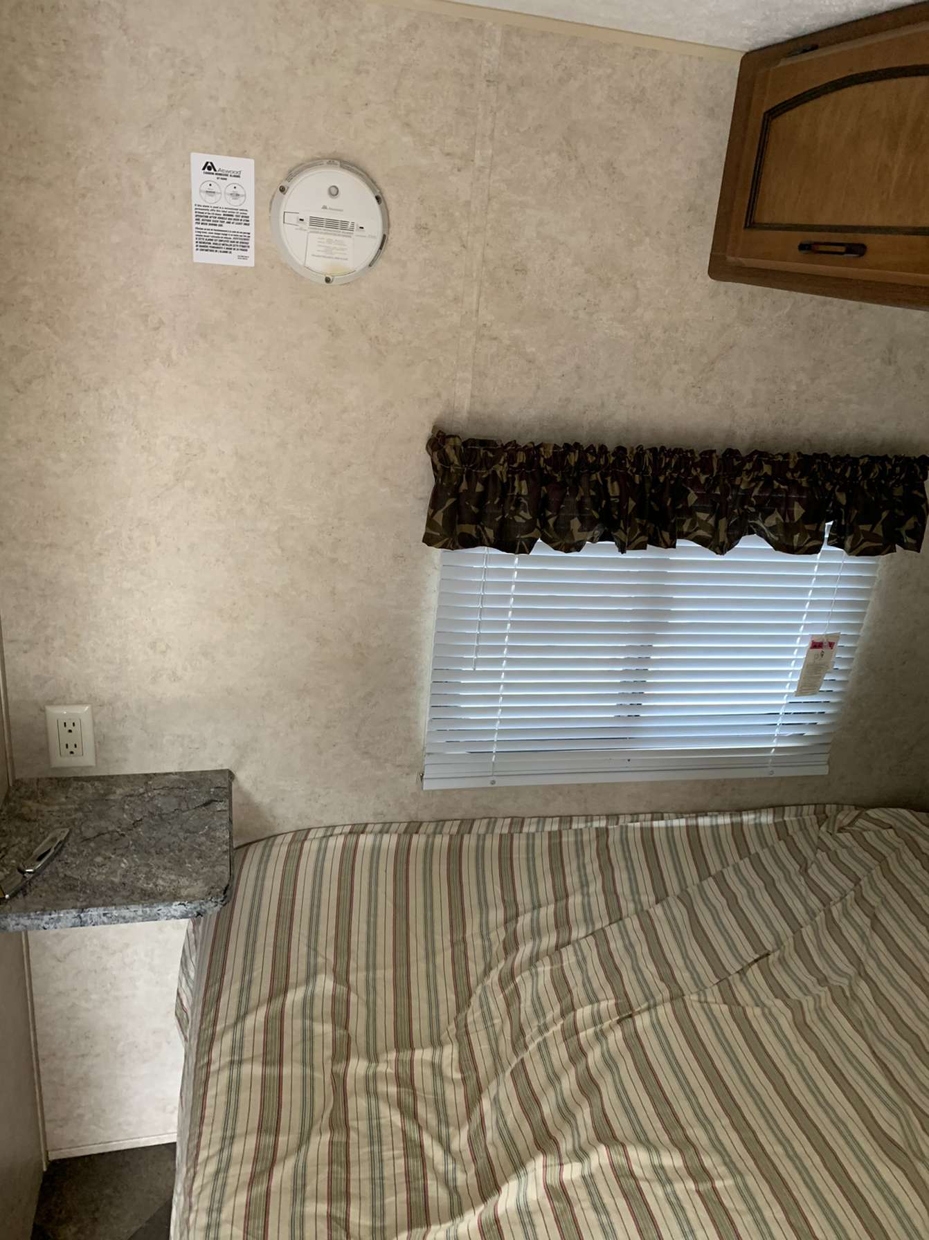 Cozy RV rental bedroom interior with striped bed, window, nightstand, smoke detector, and wooden cabinet.. Keystone RV Summerland 2013