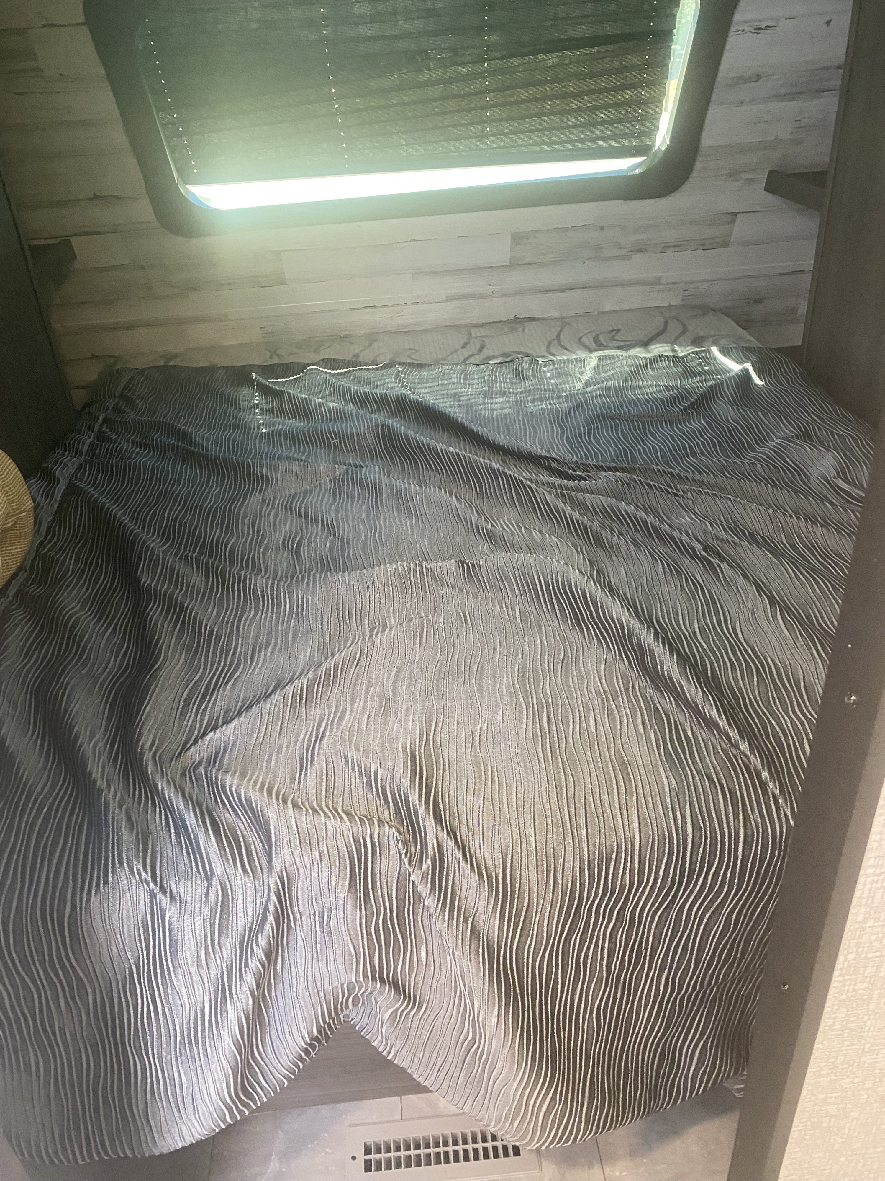 Cozy RV rental bedroom featuring a dark, textured comforter, window with blinds, and wood-paneled walls.. Dutchmen Kodiak 2021