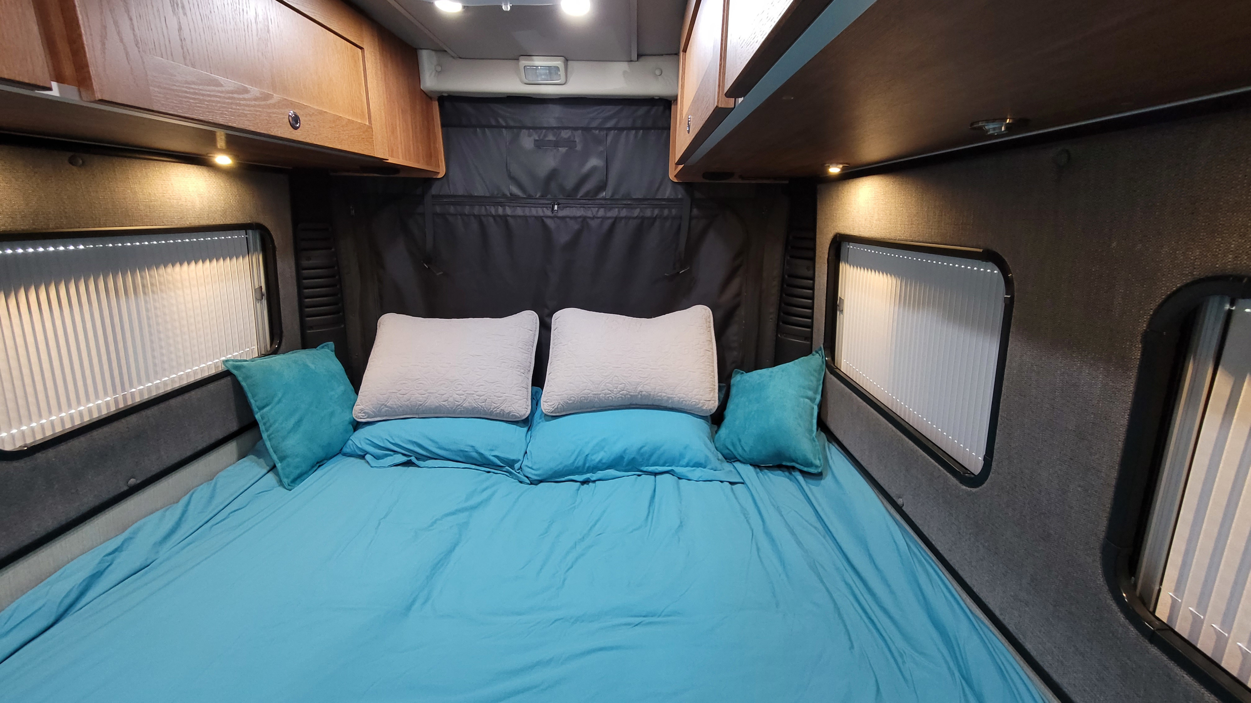 Cozy RV rental bedroom with a made bed, blue sheets, white pillows, side window blinds, and overhead wooden cabinets.. Roadtrek Play 2021