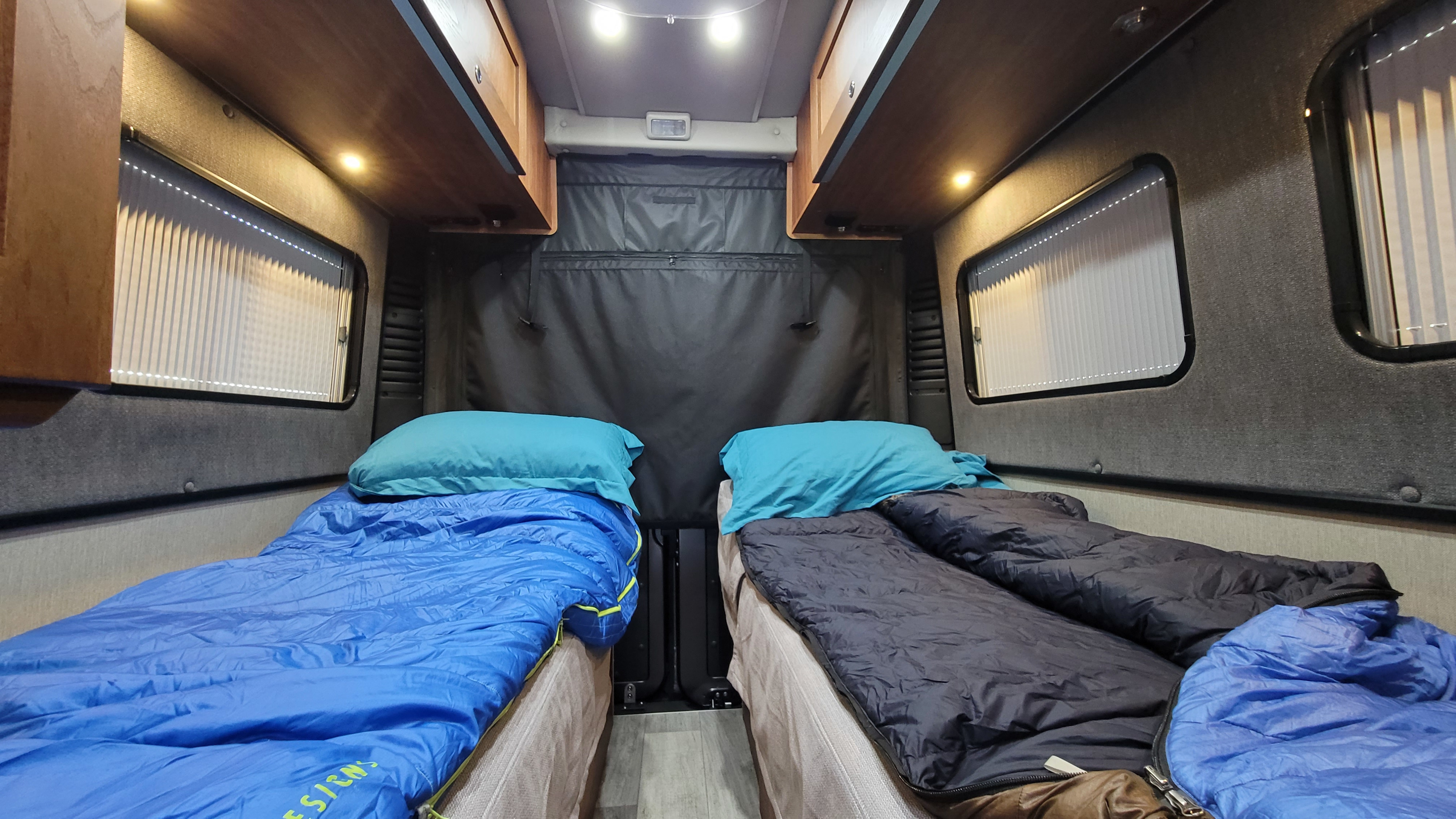 Cozy RV rental interior with two sleeping setups, overhead cabinets, and windows with blinds. Ready for adventure.. Roadtrek Play 2021