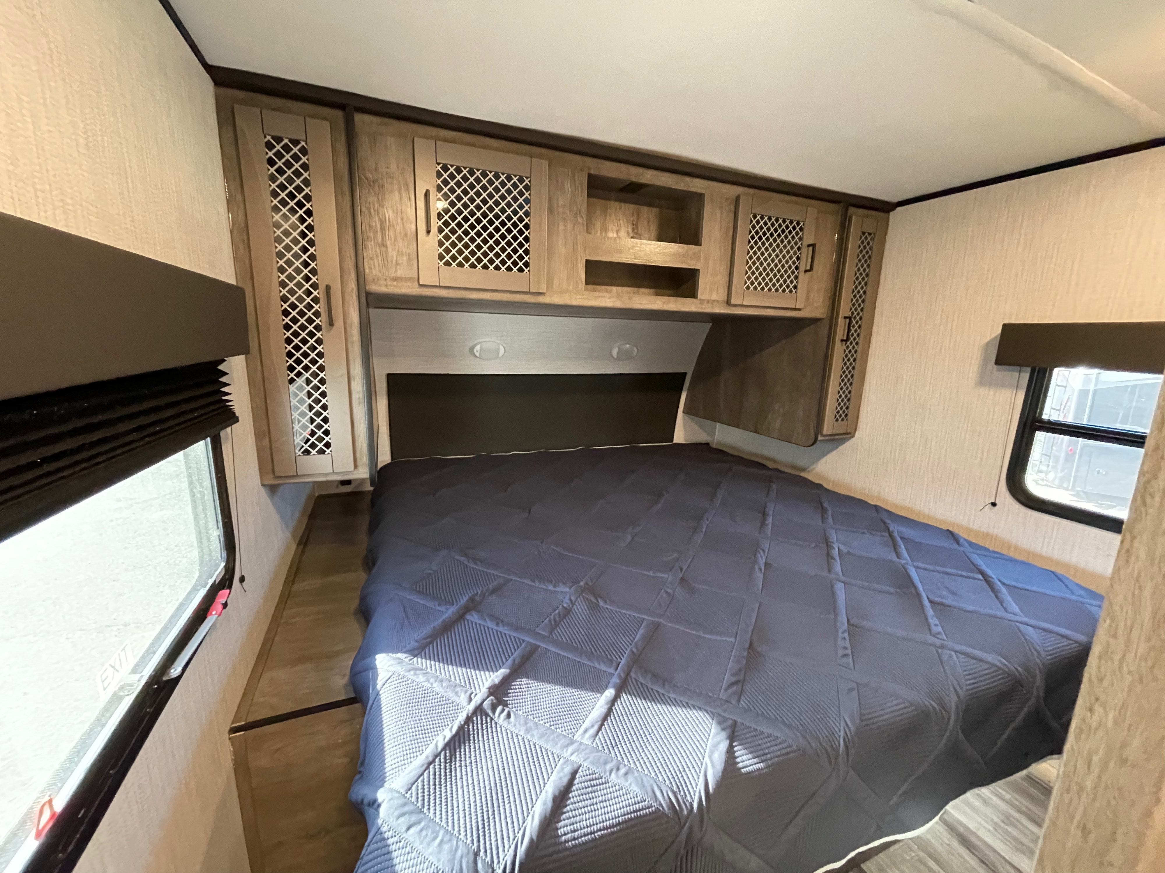 Modern RV rental bedroom features a blue quilted bed, compact overhead cabinets, and windows for a comfy travel stay.. Heartland North Trail 2021